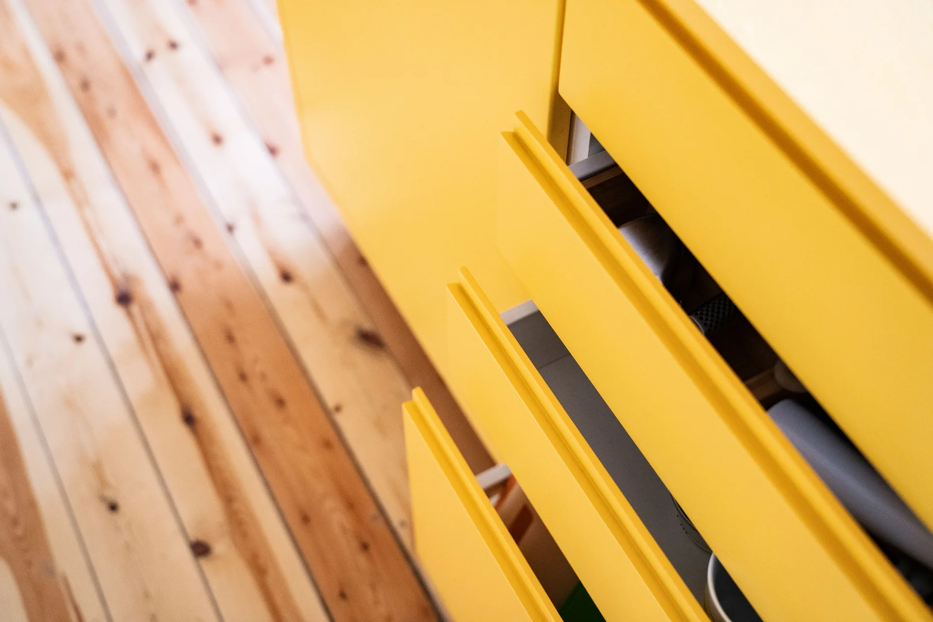Detail of open matte yellow handleless cabinet drawers installed over a light brown knotted wood plank floor.