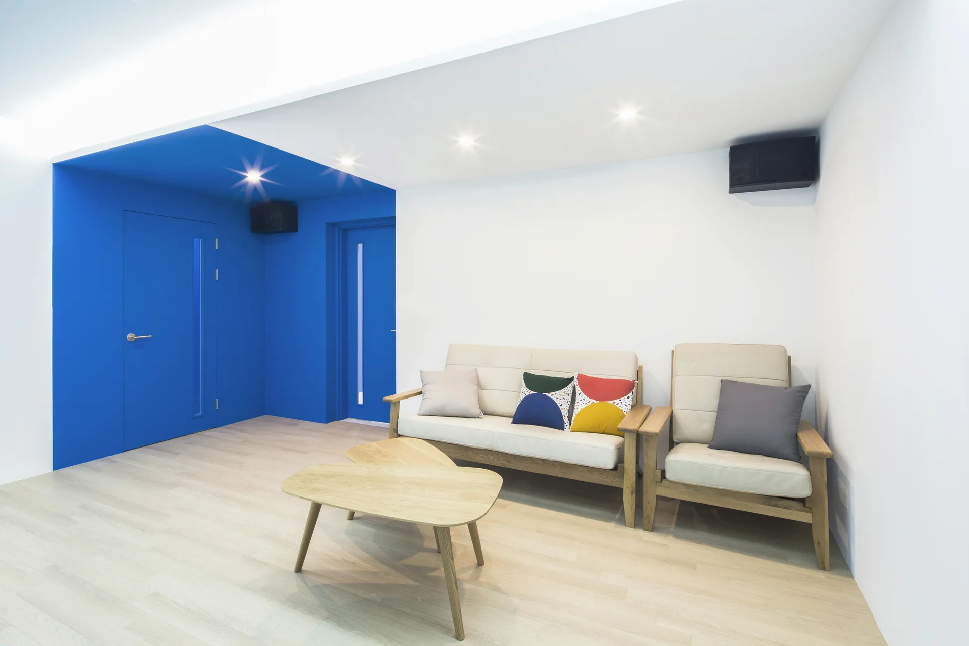 Modern living room showing a beige wood-frame sofa and armchair alongside a nesting coffee table, set against a bold blue corner alcove.