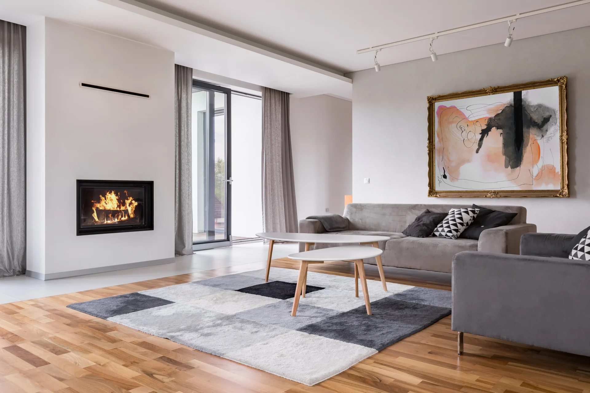 A modern living room features a built-in fireplace, grey sofa, nested coffee tables, and a large abstract painting in an ornate gold frame.