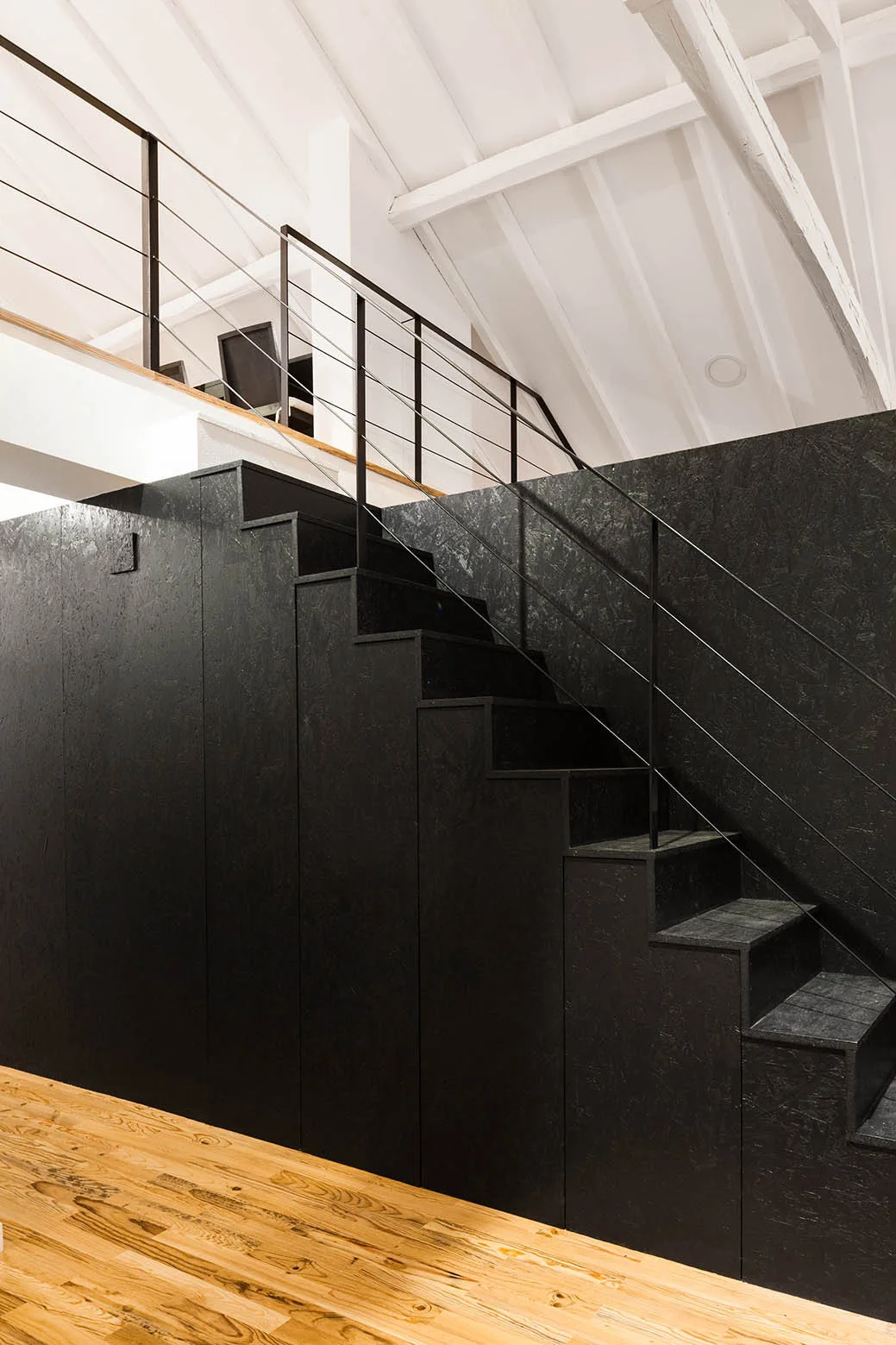 A modern black wooden staircase with a sleek cable railing sits on a light wood floor beneath a white vaulted ceiling with exposed beams.