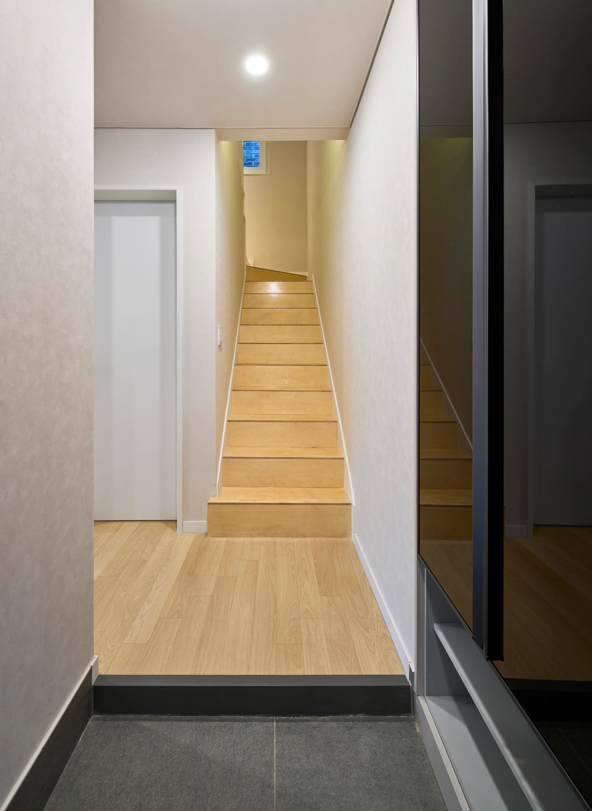 A minimalist entryway corridor featuring light wood stairs, a dark stone tile landing, and a glossy dark reflective cabinet panel.