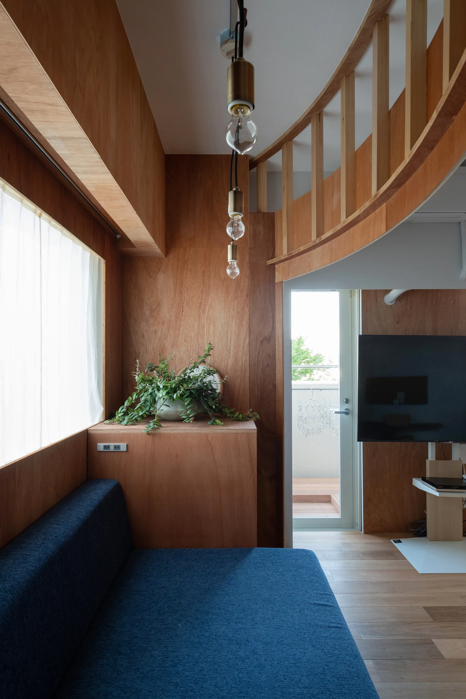 A compact modern living room featuring warm plywood paneled walls, a deep blue sofa, three hanging bulb pendant lights, and a curved wooden loft railing.