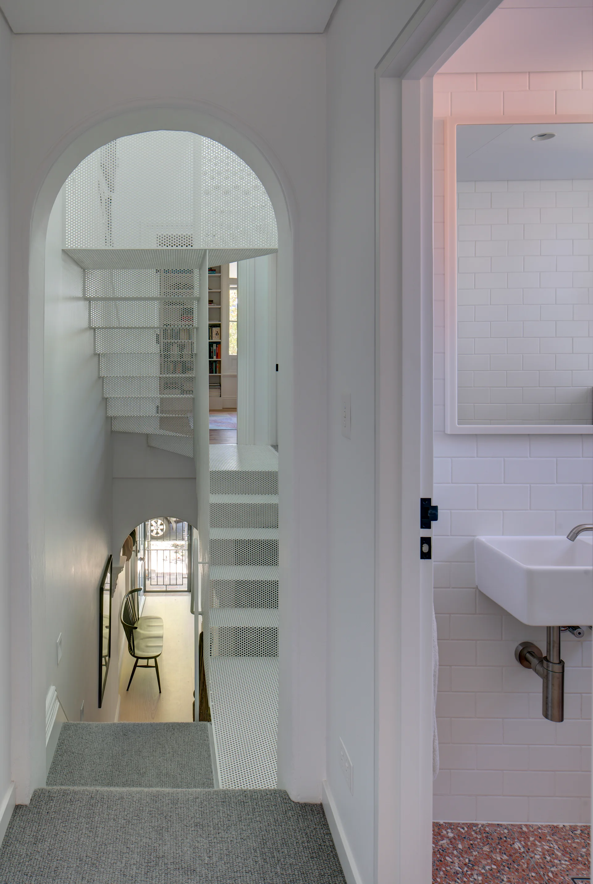 A view through a white archway reveals a perforated metal staircase next to a bathroom with subway tiles and terrazzo flooring.