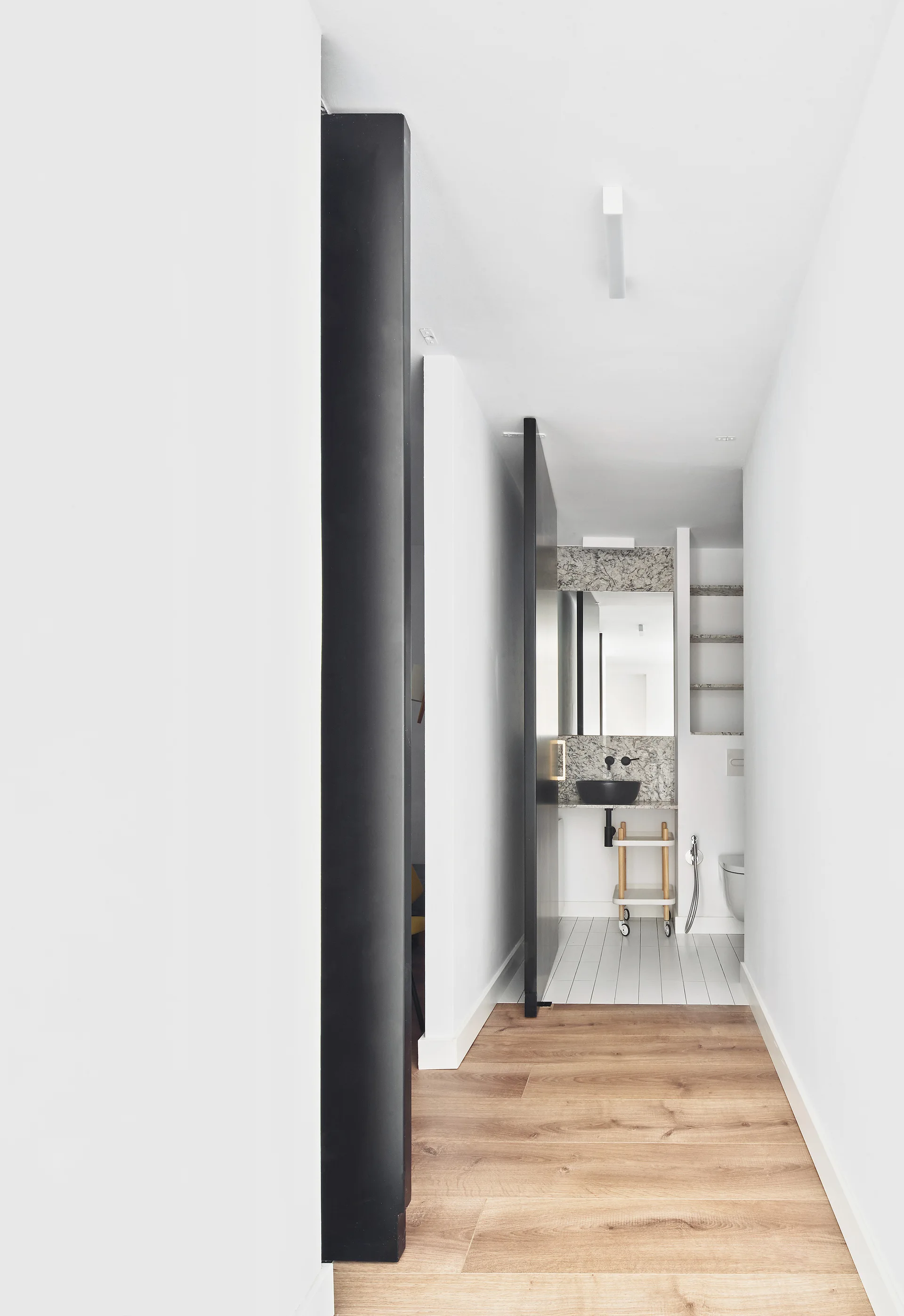 A narrow hallway leads into a minimalist white bathroom featuring a black sliding door, a marble backsplash vanity, and wood flooring.