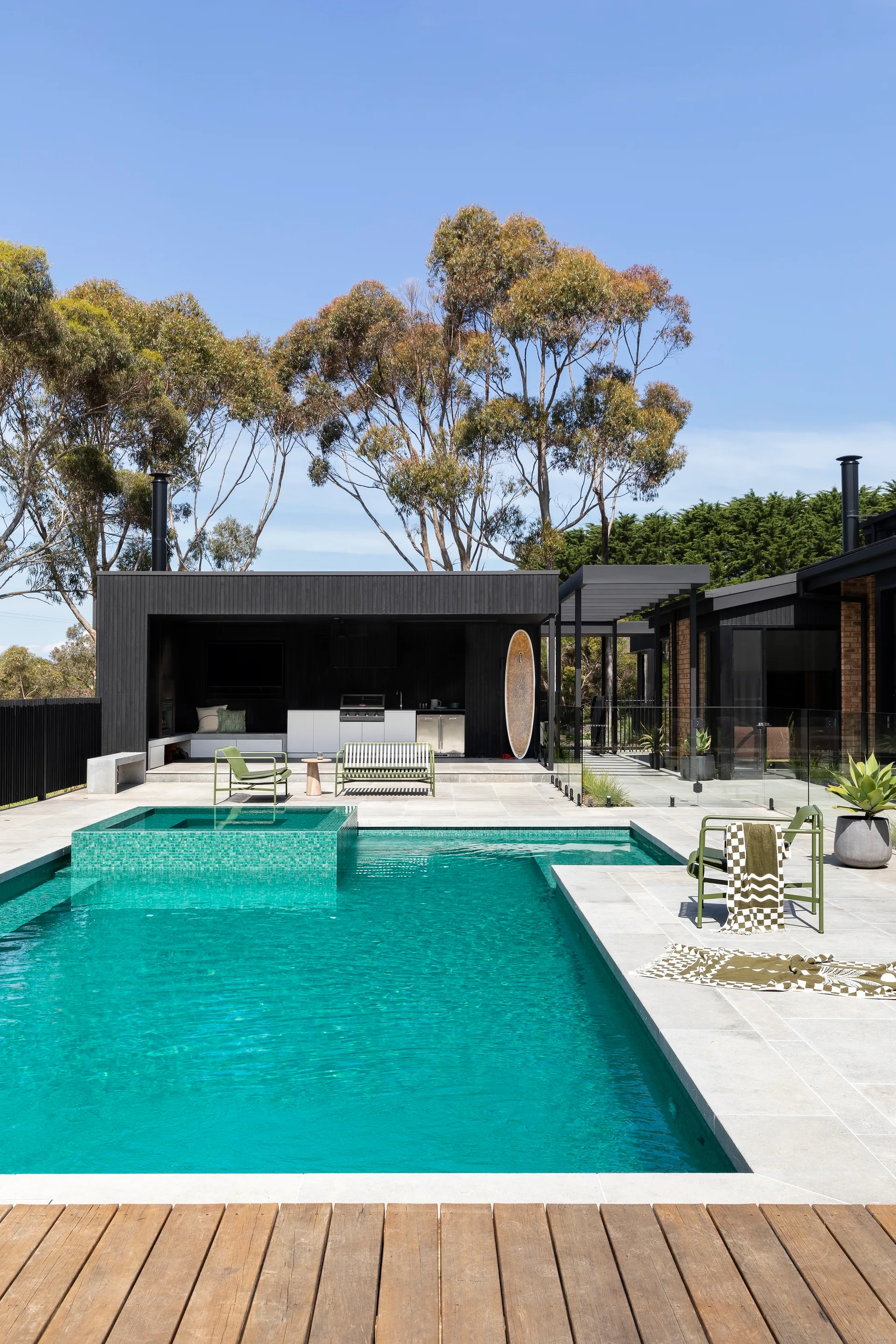 A modern turquoise swimming pool is surrounded by grey stone pavers, olive green chairs, and a black wood pool house with an outdoor kitchen.