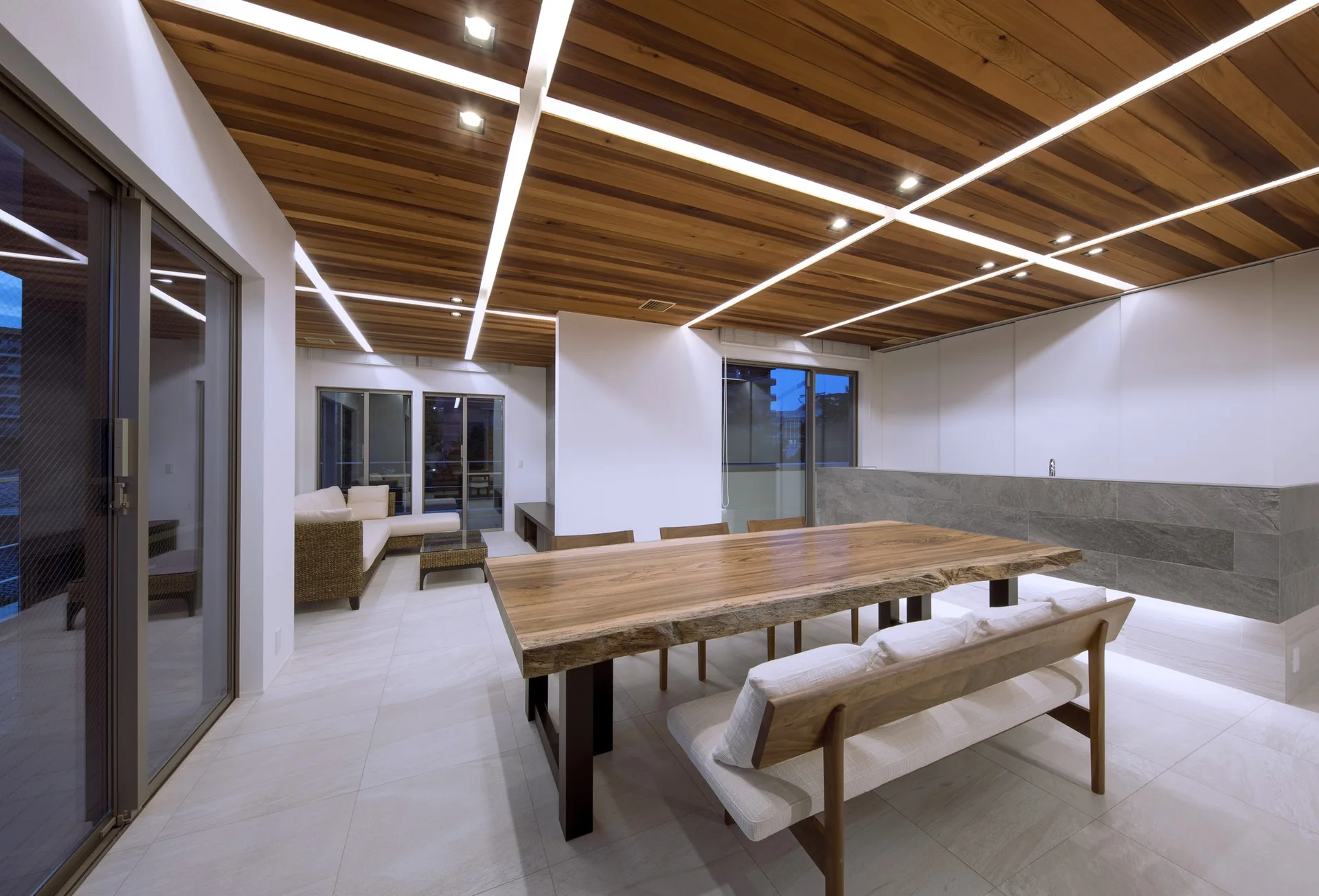 A modern dining area features a large live-edge wooden table, a matching bench, a slatted wood ceiling with linear light strips, and tile flooring.