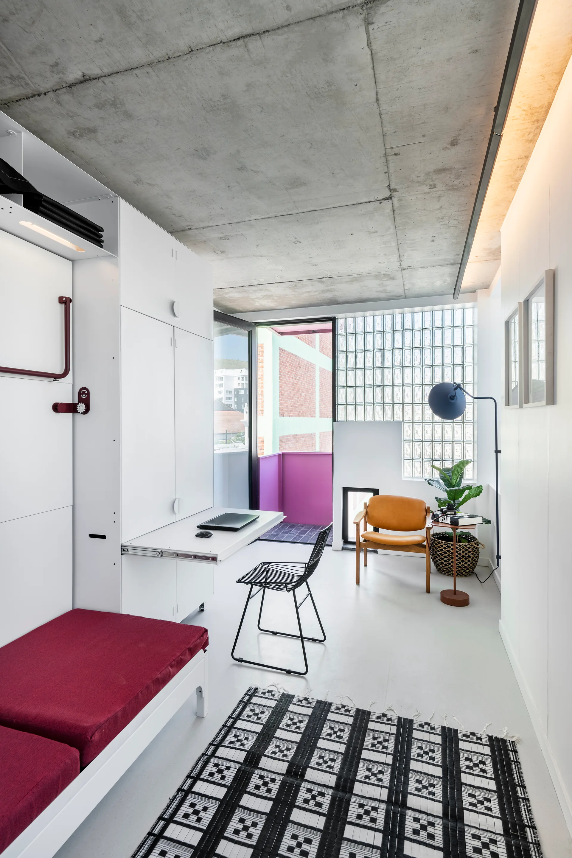 A modern studio apartment features a foldaway desk, a tan armchair, an exposed concrete ceiling, and a translucent glass block window wall.