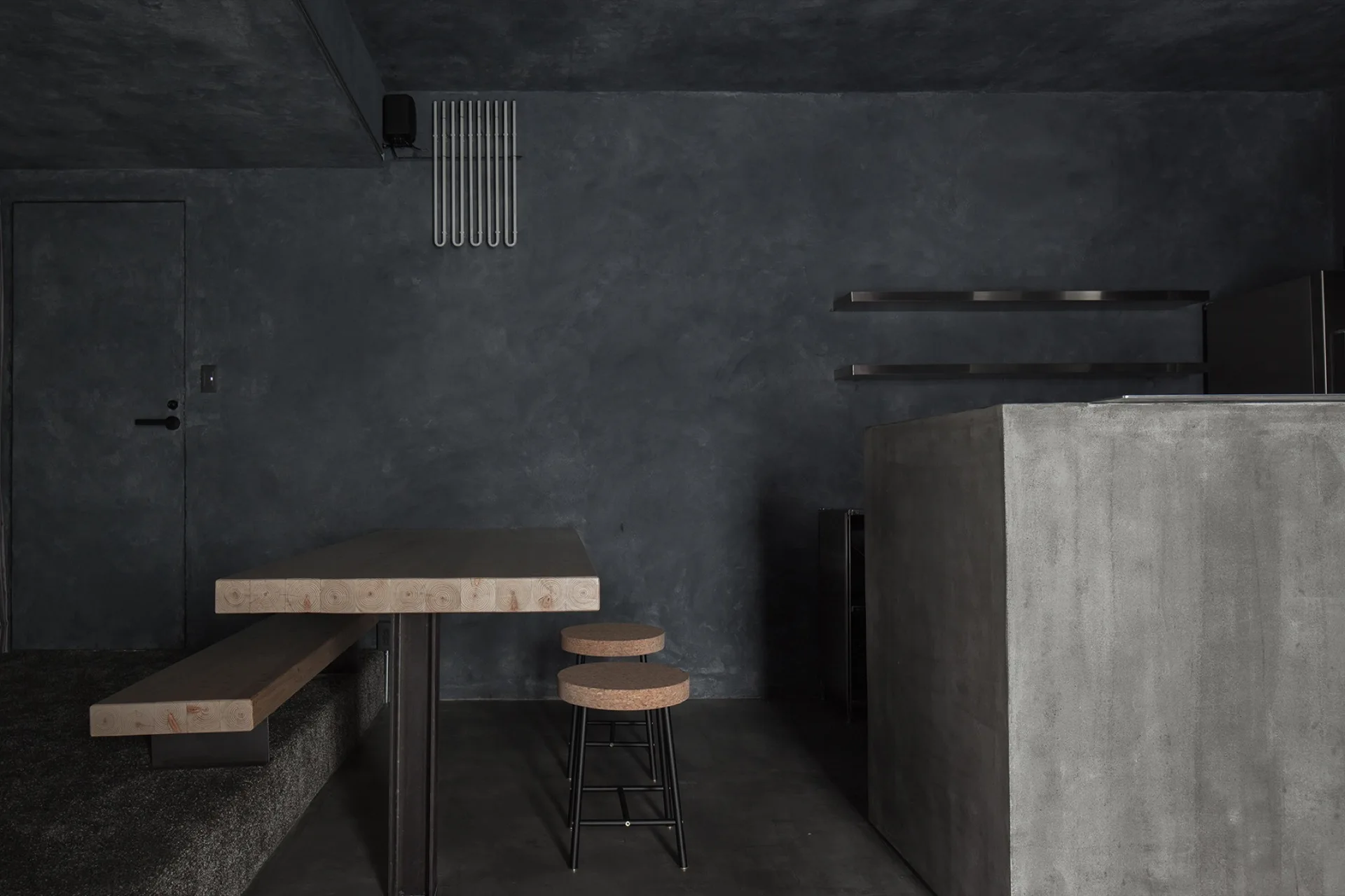 A dark minimalist dining area featuring charcoal textured walls, a thick wood slab table with a bench, cork stools, and a concrete island counter.