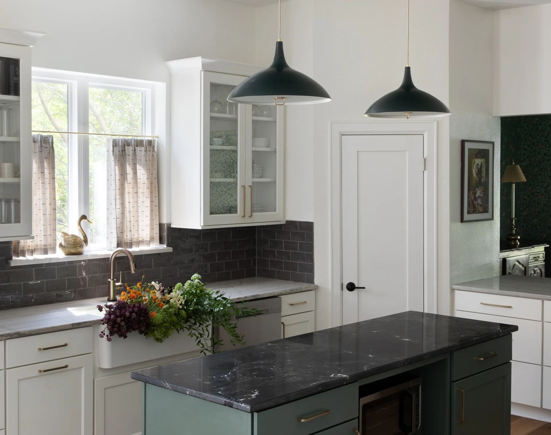 A kitchen features a dark green island with a black marbled countertop beneath matching pendant lights, white perimeter cabinets, and a dark tile backsplash.
