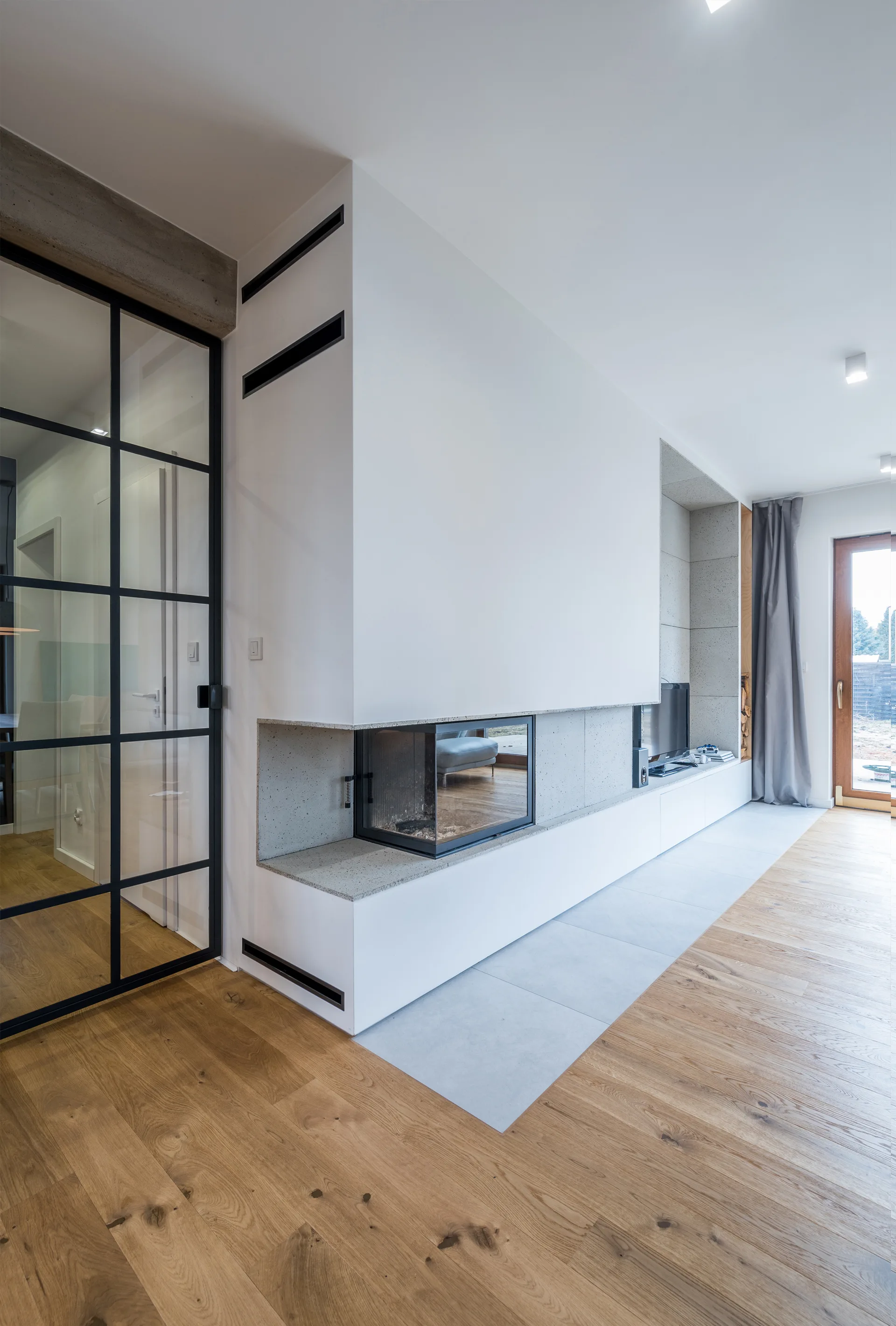 A modern living room featuring a built-in corner fireplace, a long low media console, light wood floors, and a black-framed glass wall.