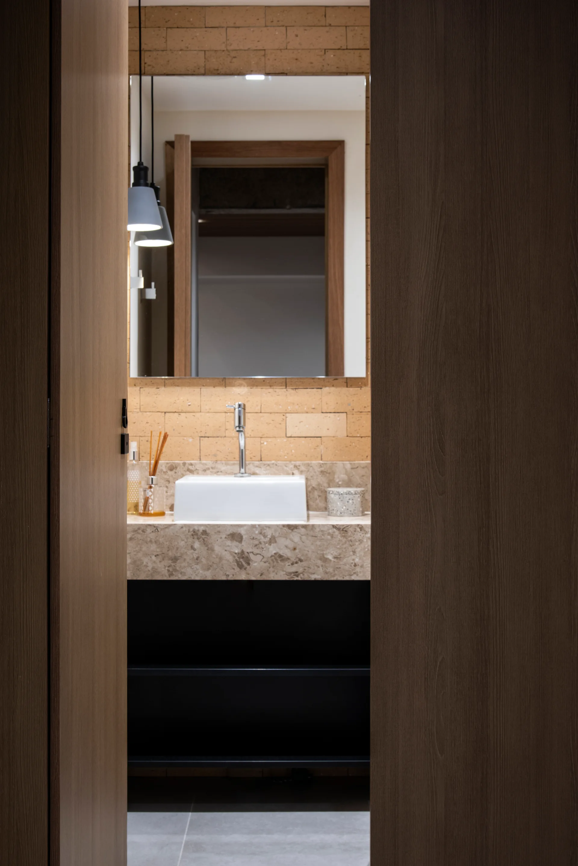 A modern bathroom vanity with a white vessel sink on a veined stone countertop against a light brown brick wall, viewed through a wood doorway.