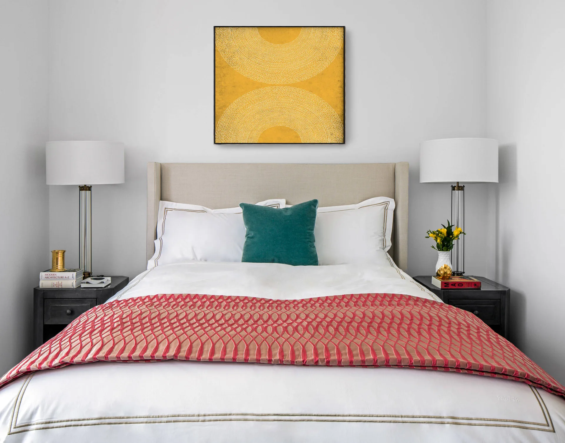 A symmetrical bedroom features a beige upholstered bed, white lamps on black nightstands, a central teal pillow, and yellow framed wall art.