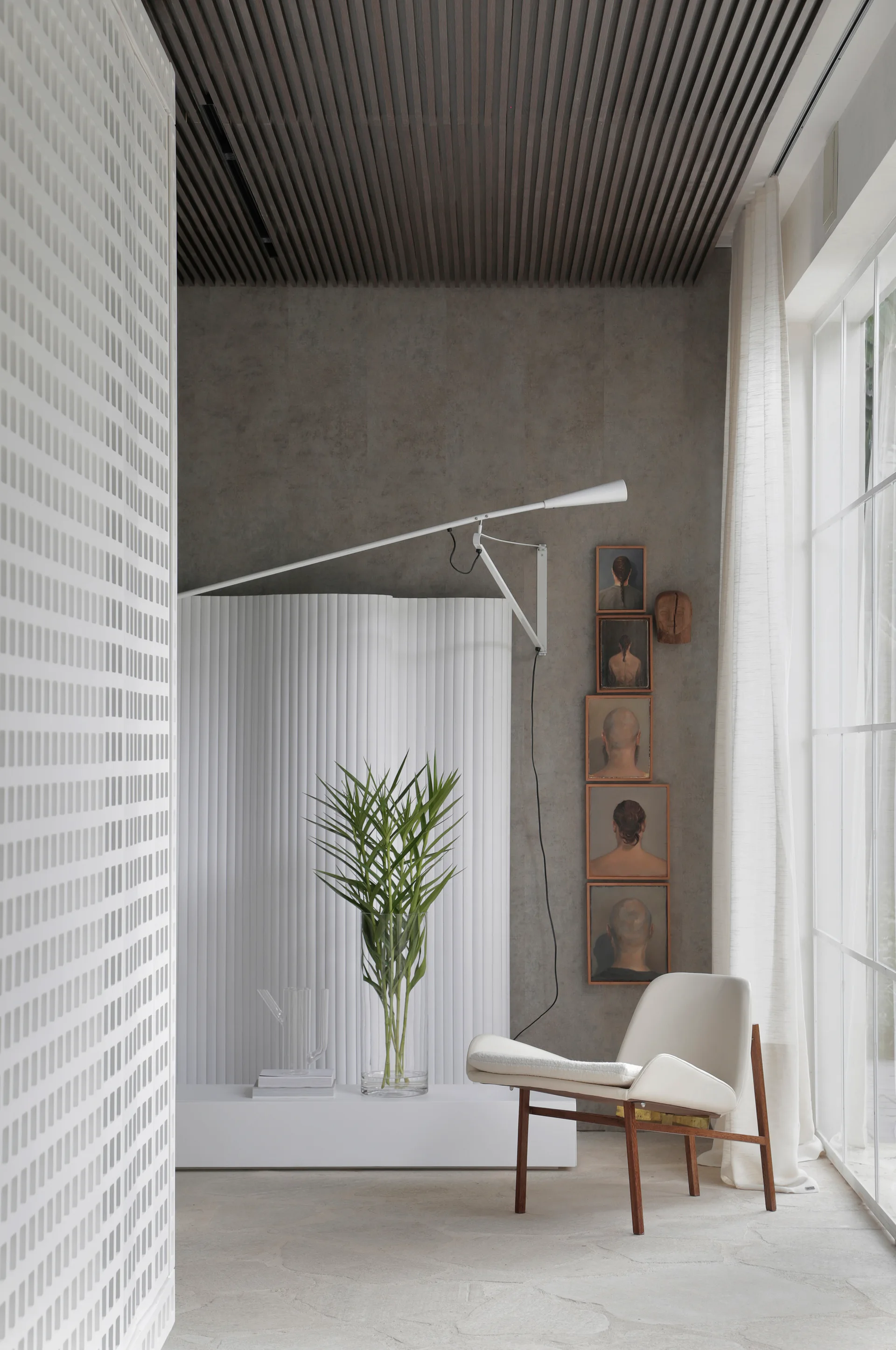 A minimalist lounge area features a white armchair beside a grid window, a fluted partition wall, and a dark slatted wood ceiling.