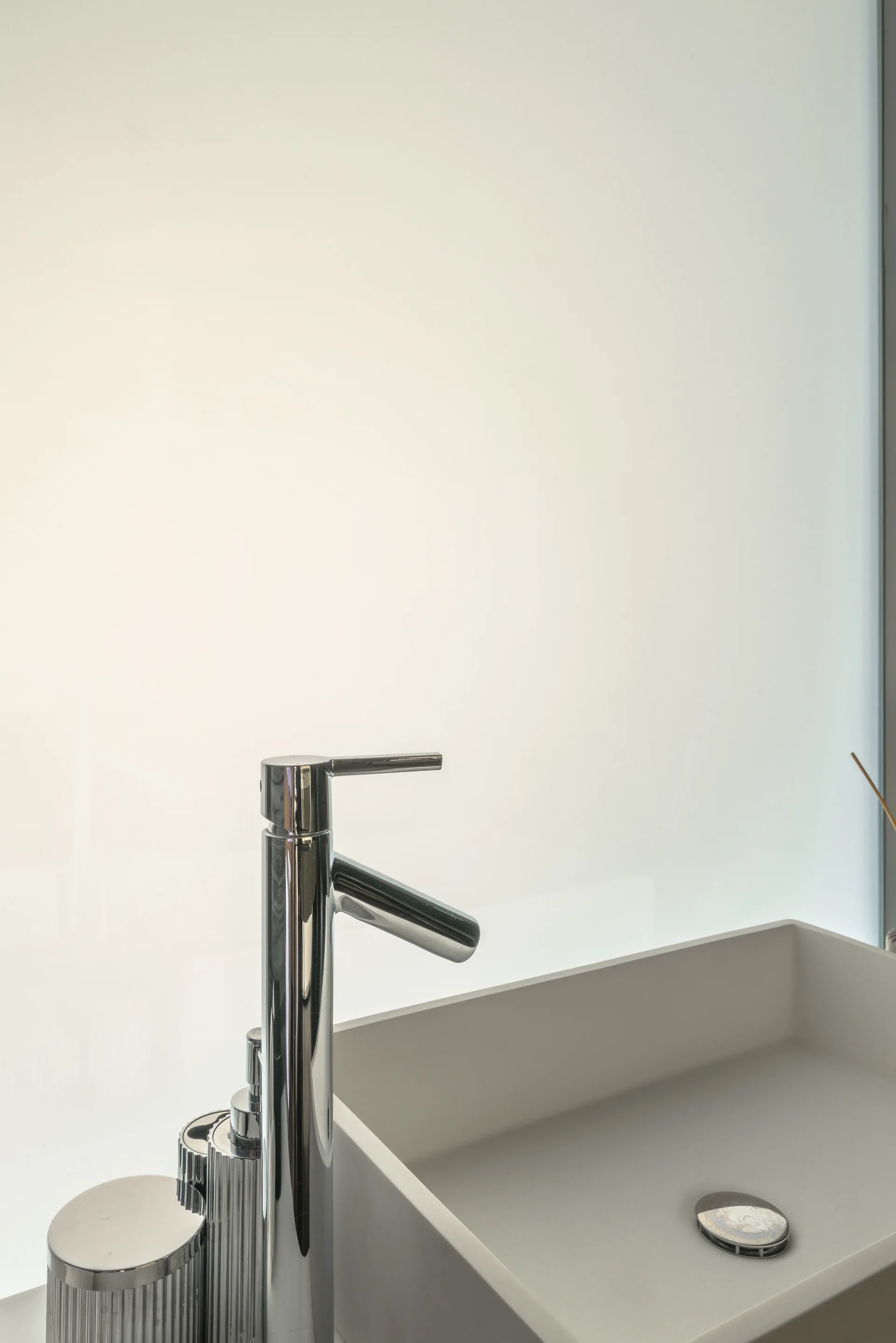 A close-up of a tall polished chrome faucet and a matte white vessel sink set against a backlit frosted glass background.