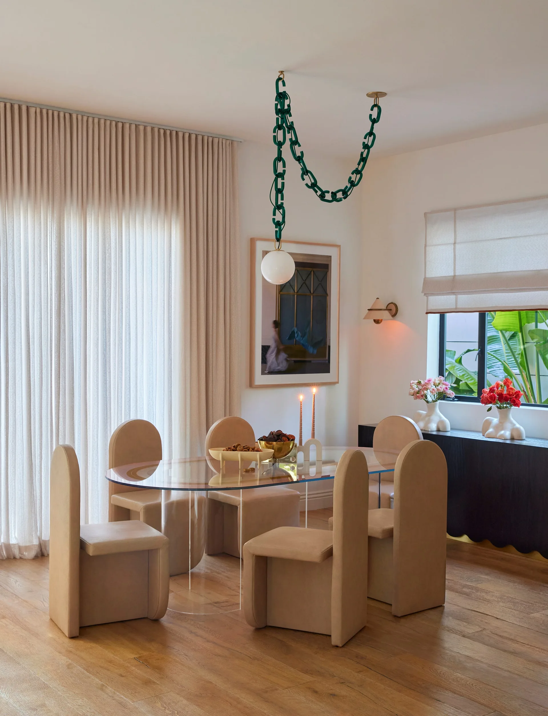 A modern dining room featuring tan arched-back chairs around an oval glass table, with a green chain-link pendant light hanging above.
