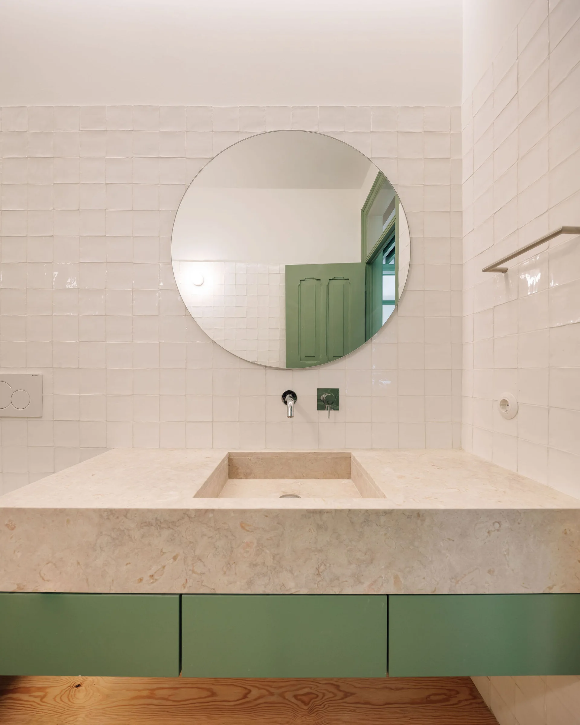 A minimalist bathroom features a stone floating vanity with sage green cabinets, a circular mirror, and glossy white square wall tiles.