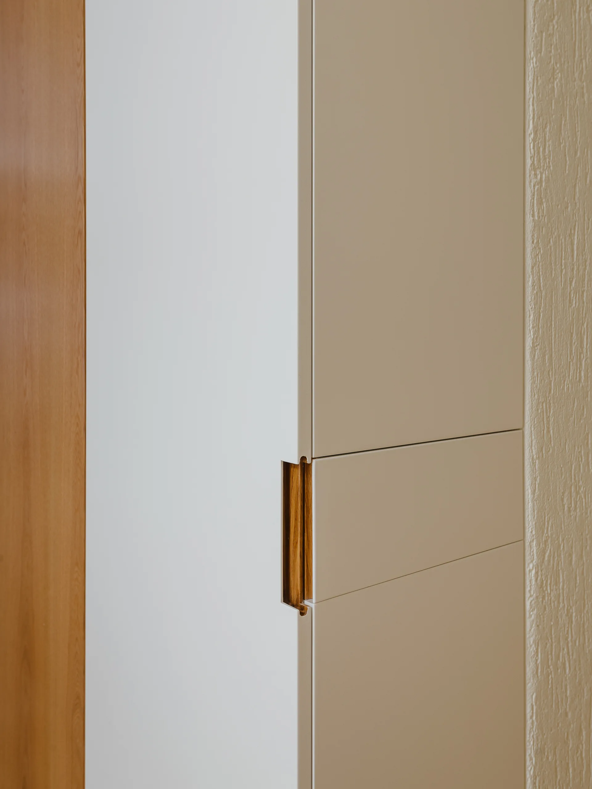 A close-up of matte white and beige cabinet panels featuring a custom recessed vertical handle lined with natural wood veneer.