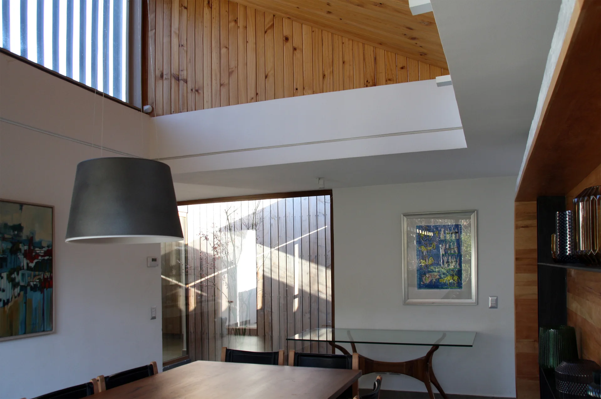 A dining room features an angled wood plank ceiling, a dark drum pendant light, a wooden table, and a glass-topped console table against a white wall.