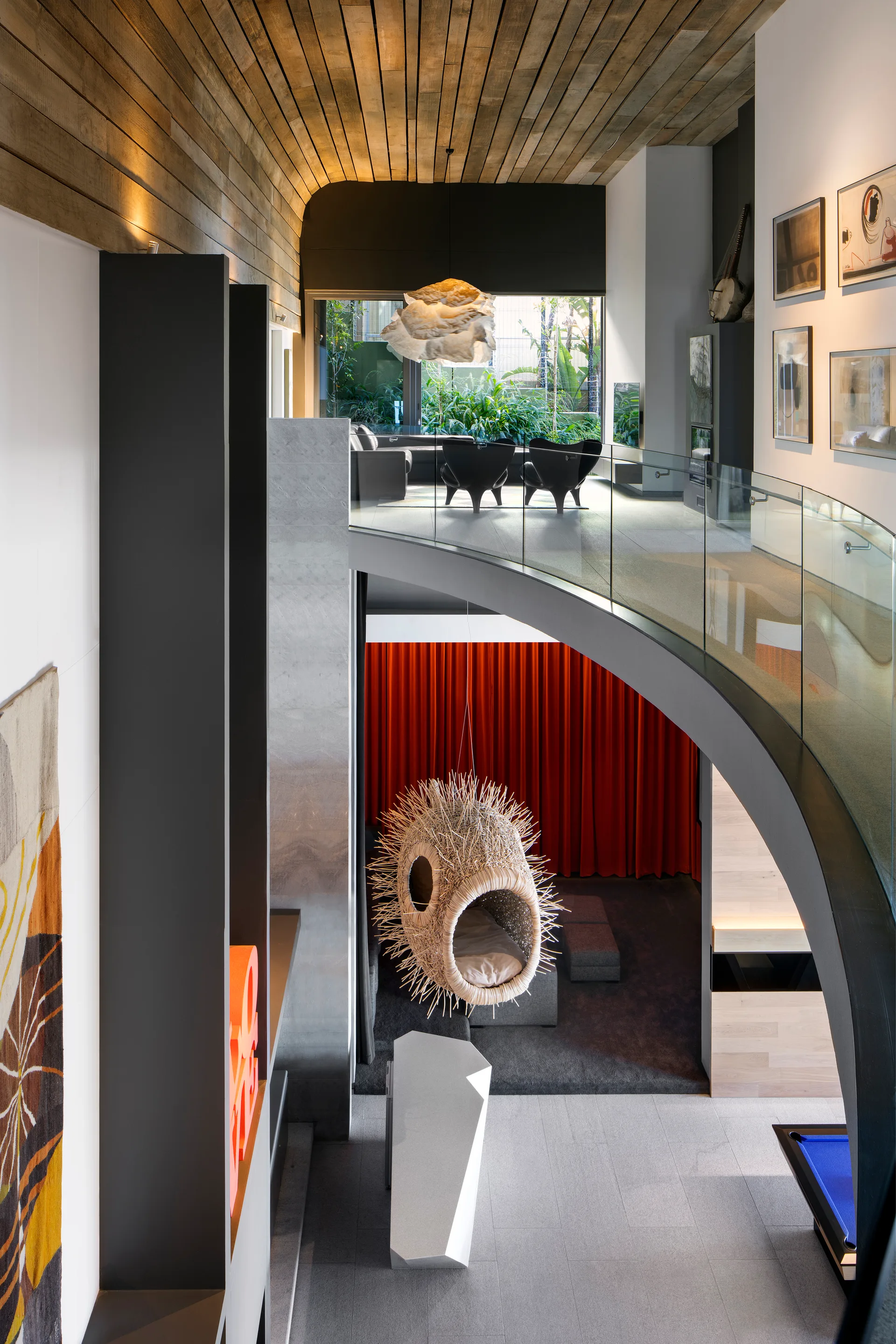 A modern multi-level home interior featuring a curved staircase with a glass railing, a wood plank ceiling, and a hanging spiky pod chair.