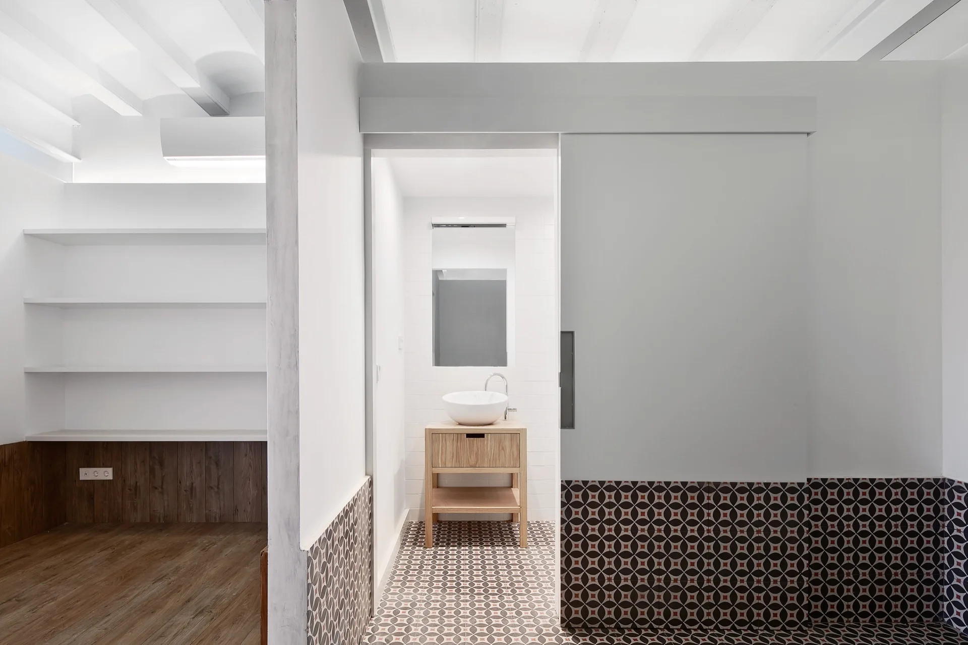 A minimalist bathroom with a light wood vanity, white vessel sink, geometric tile floor, and a large white sliding pocket door.