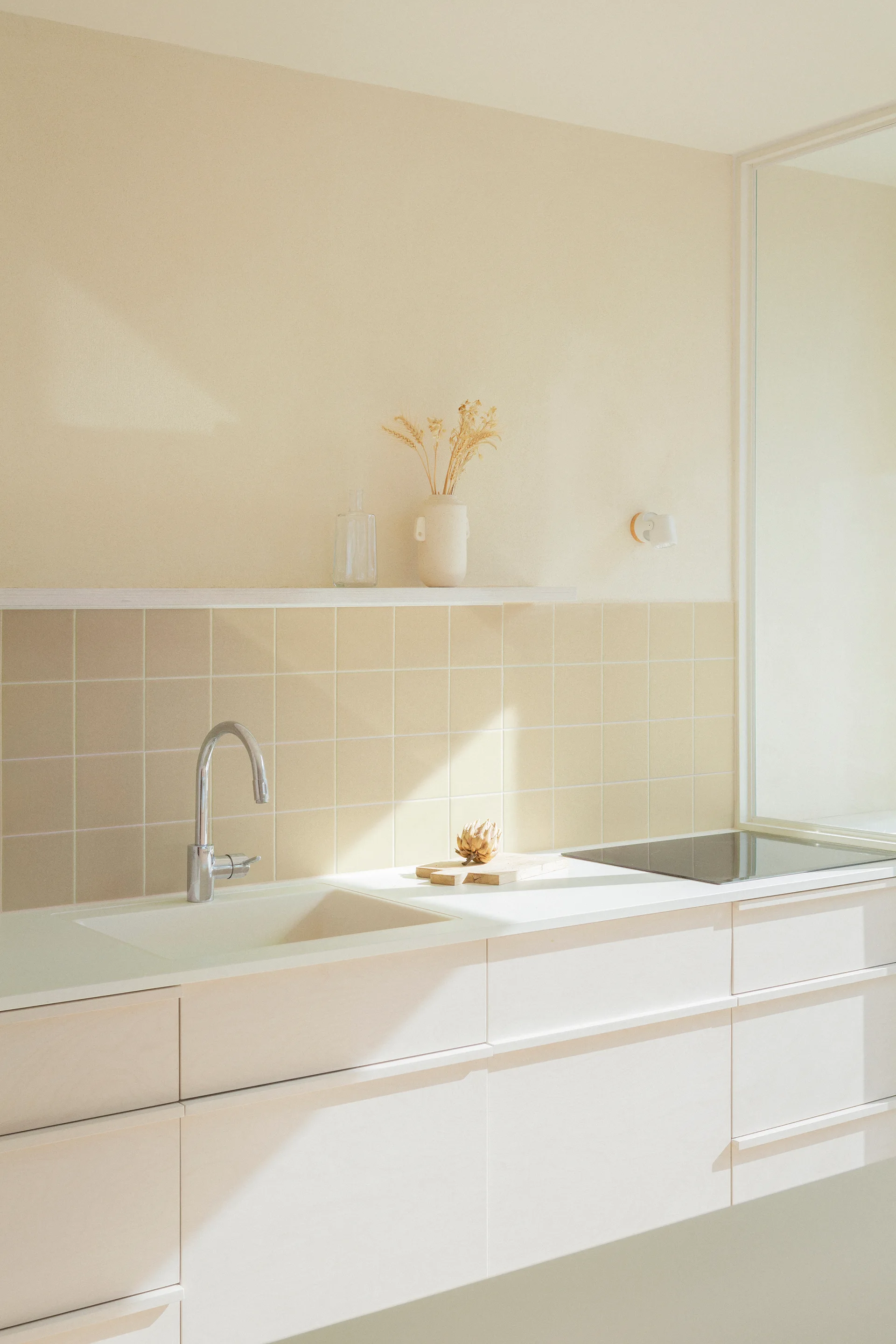 A minimalist kitchen features cream cabinets, an integrated sink with a chrome faucet, and a light beige square-tiled backsplash beneath a slim shelf.