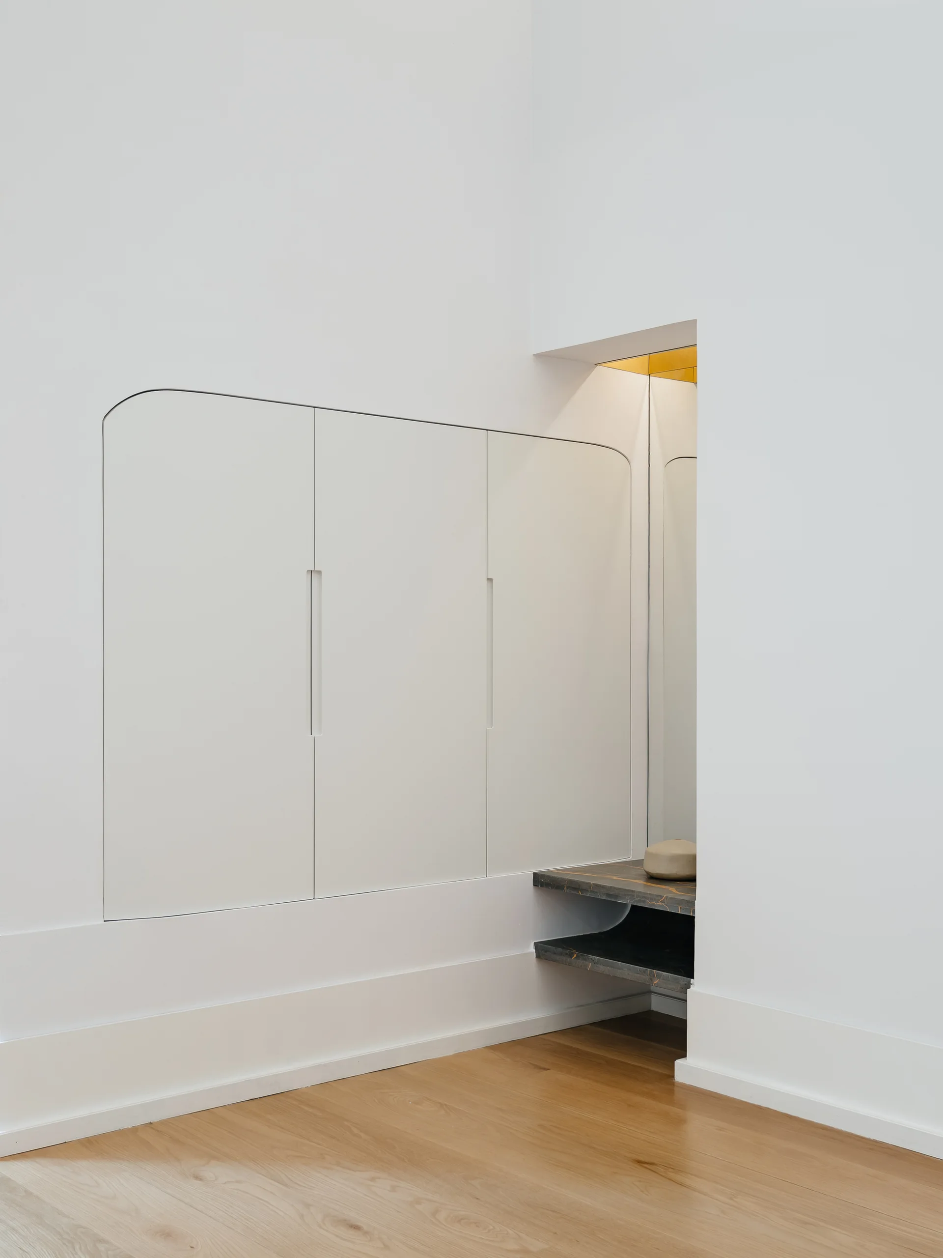 Minimalist white built-in cabinets with rounded corners stand beside an integrated niche featuring a dark veined floating shelf and warm gold lighting.
