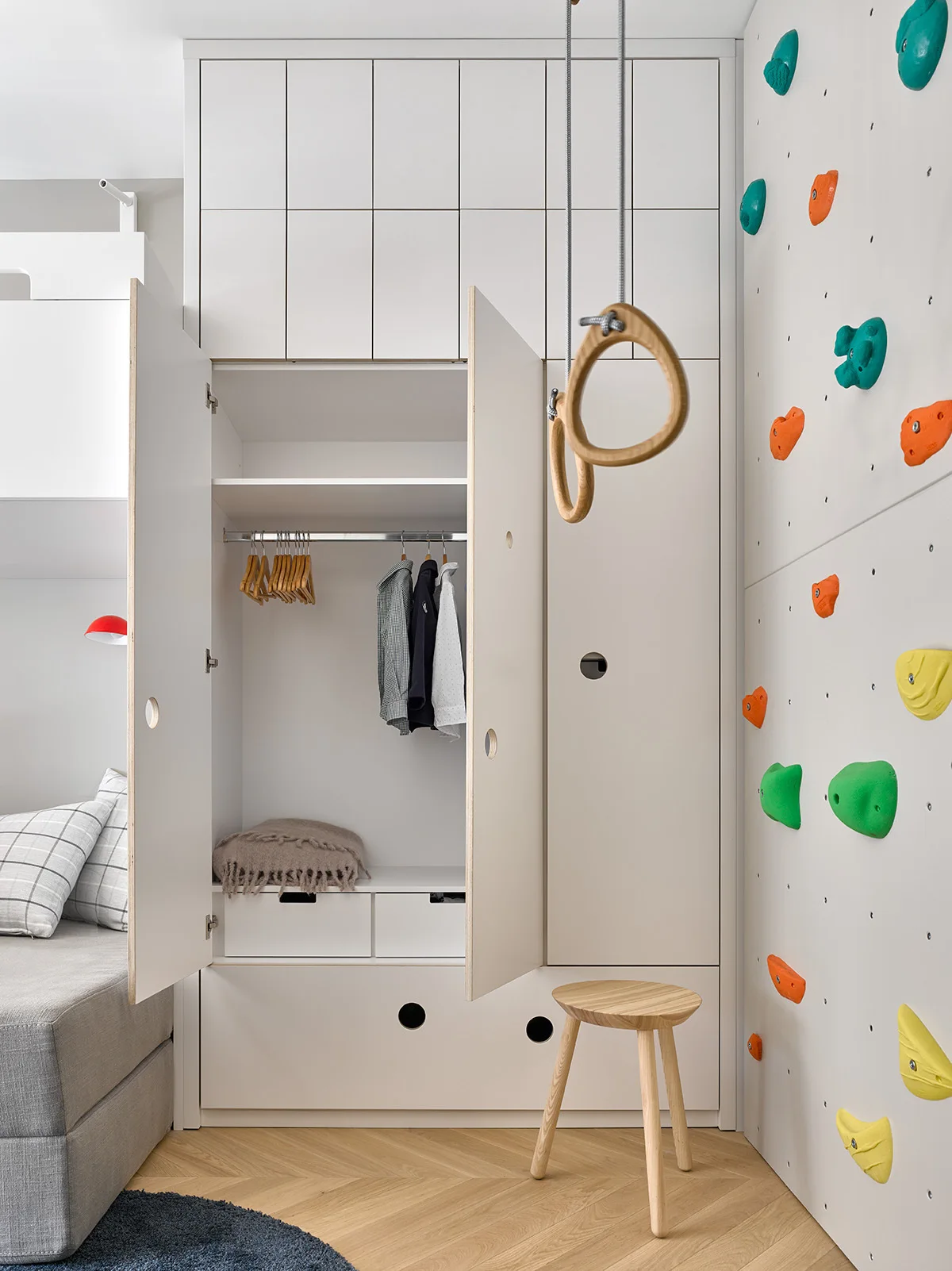 A modern children's bedroom features a built-in white wardrobe with open doors next to a climbing wall with colorful holds and hanging gymnastic rings.