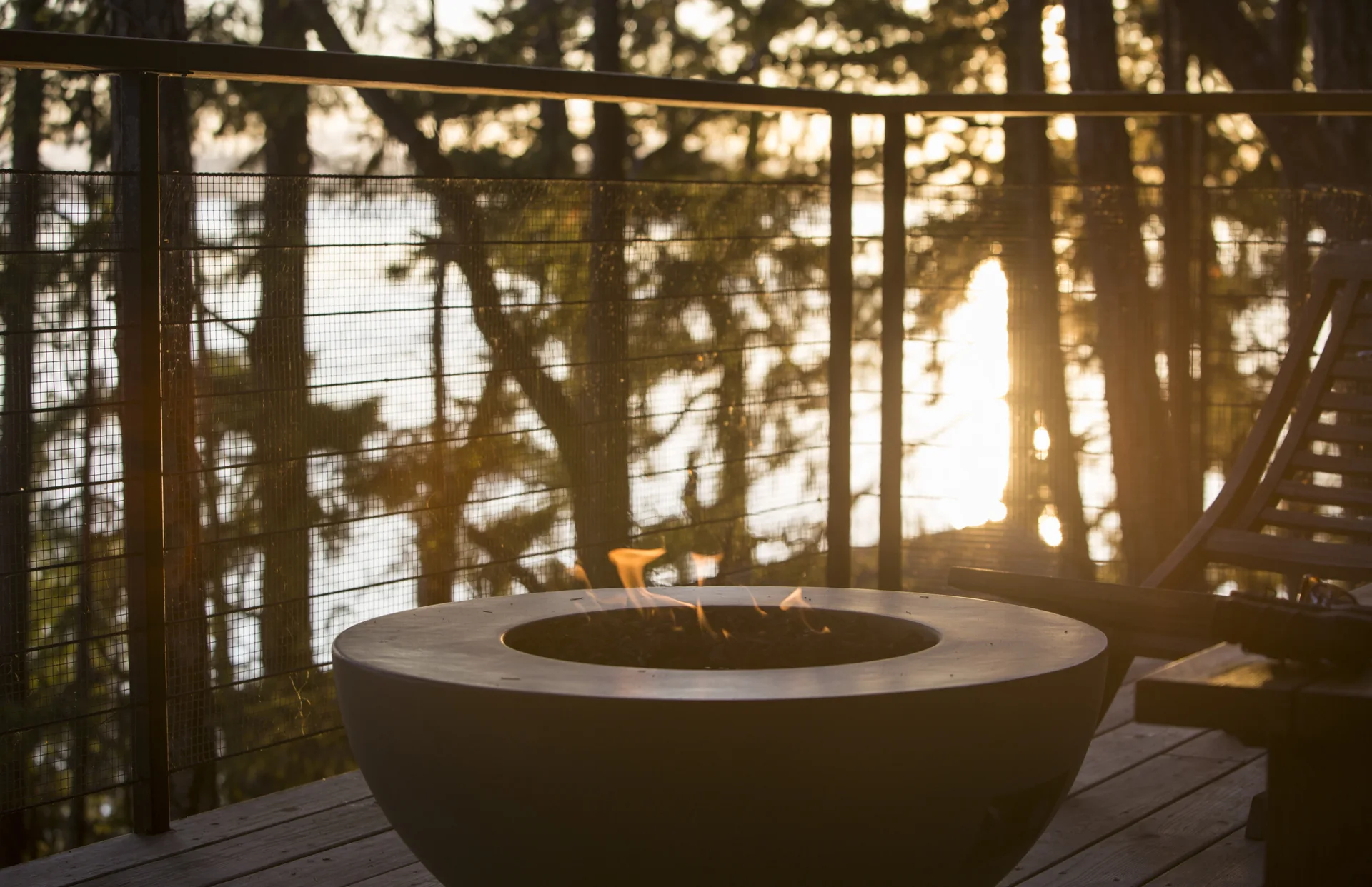 A round dark grey fire pit with burning flames sits on a wooden deck next to a wire railing overlooking a sunlit forest.