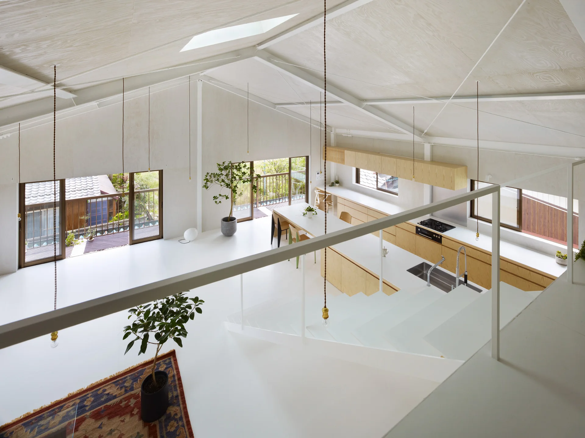 A minimalist open kitchen and dining area with light wood cabinetry, white floors, and a vaulted plywood ceiling.