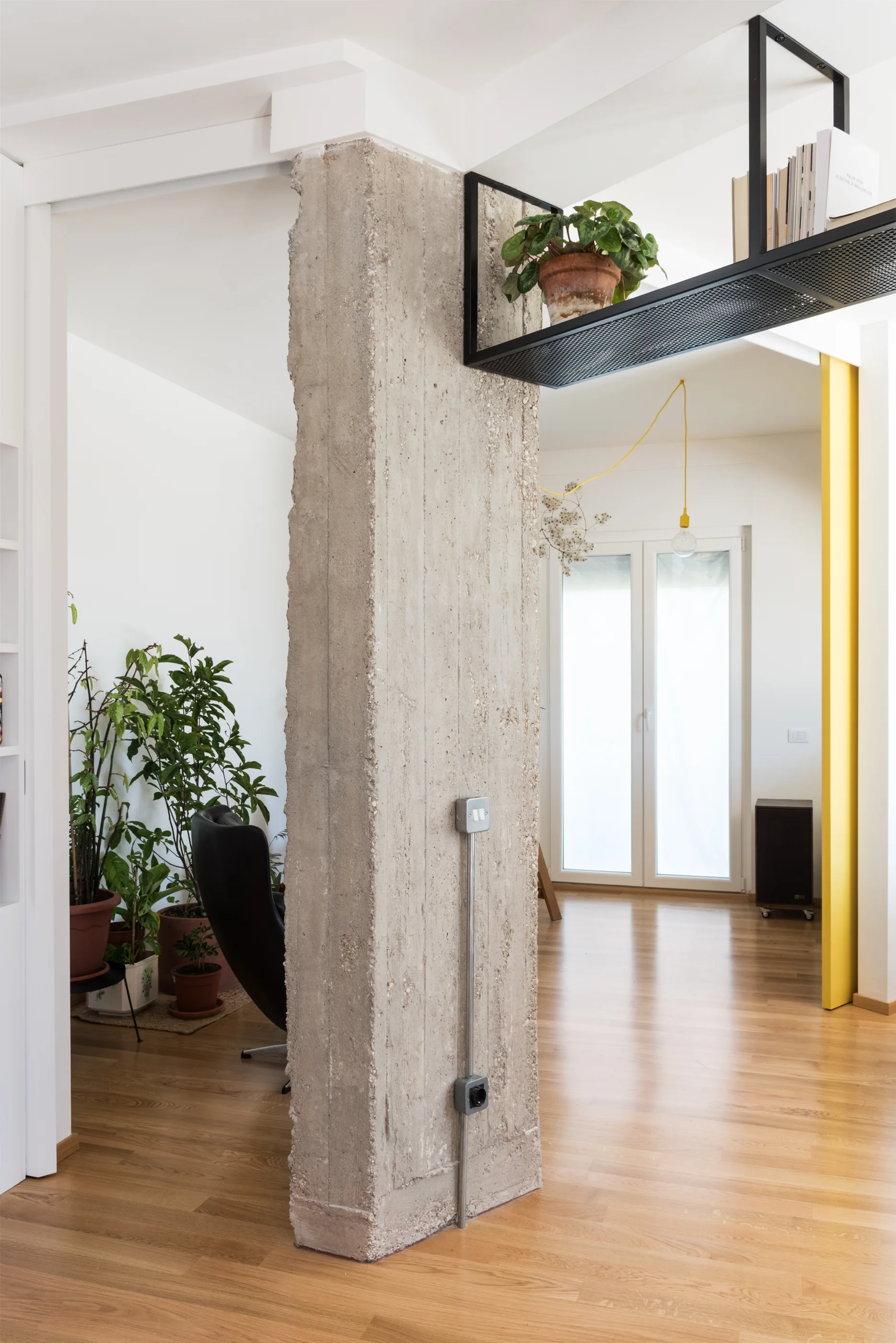 An interior room featuring an exposed raw concrete pillar, light wood floors, and a black metal suspended ceiling shelf holding plants.