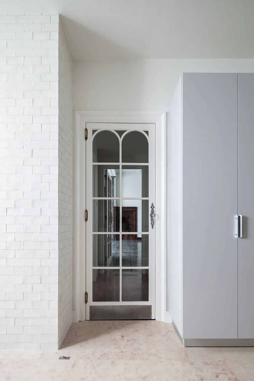 A white arched glass-paneled door is framed by a painted white brick wall and a modern light grey storage cabinet on a veined stone floor.