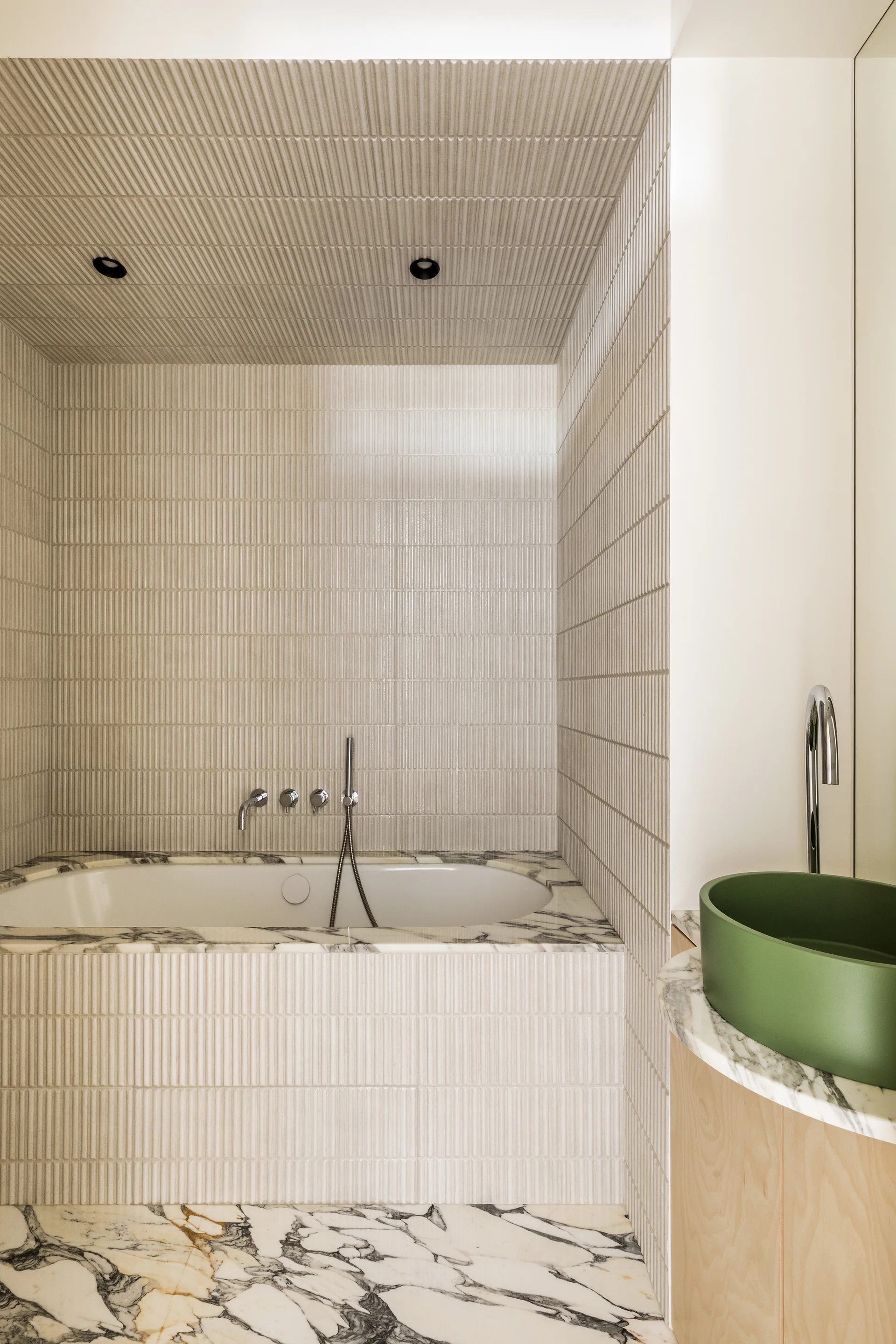 A modern bathroom features a bathtub enclosed in beige fluted tile and veined marble alongside a curved wood vanity with a matte green vessel sink.