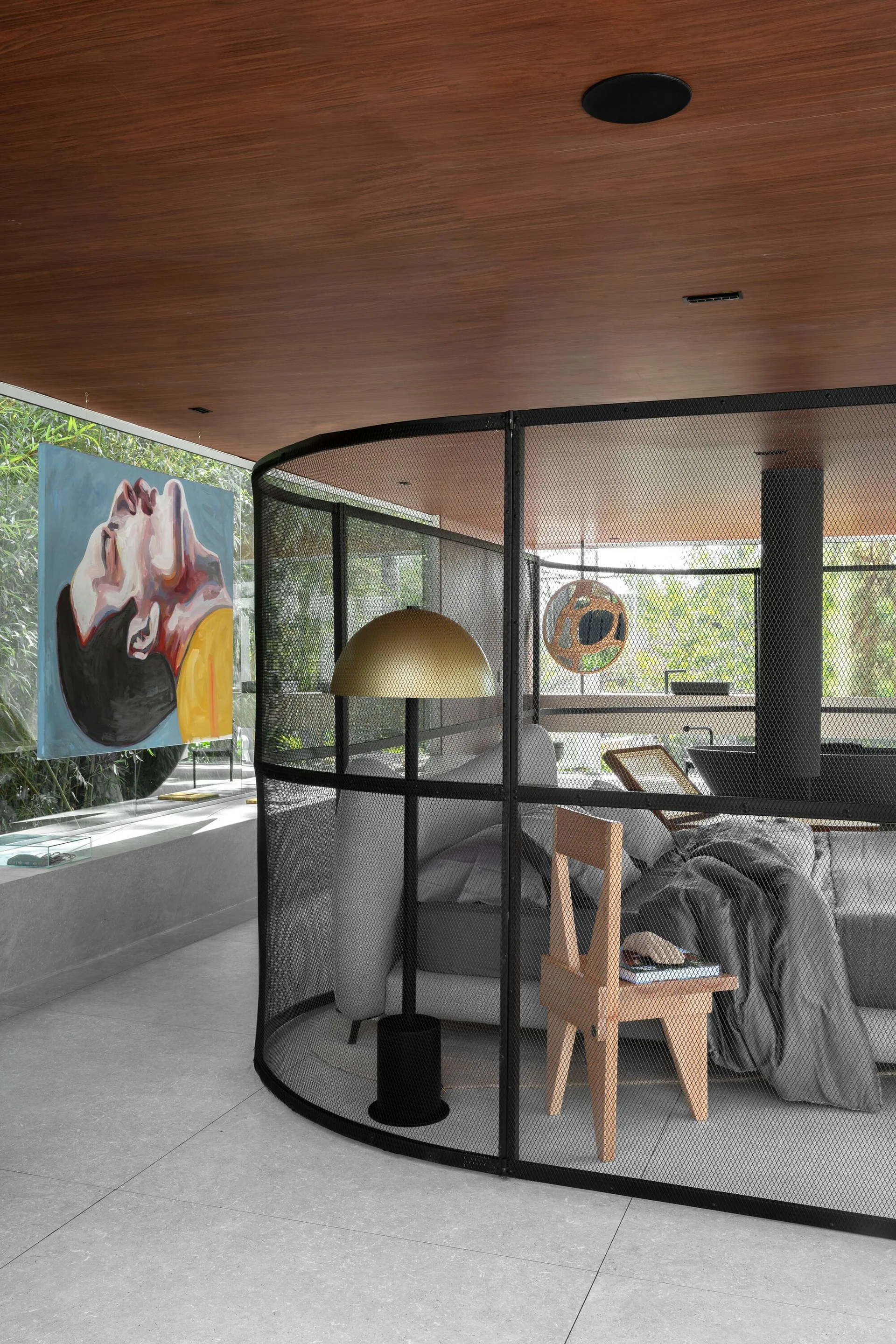 A contemporary bedroom features a curved black metal mesh screen enclosure, grey bedding, a wooden chair, and a gold dome floor lamp beneath a wood ceiling.