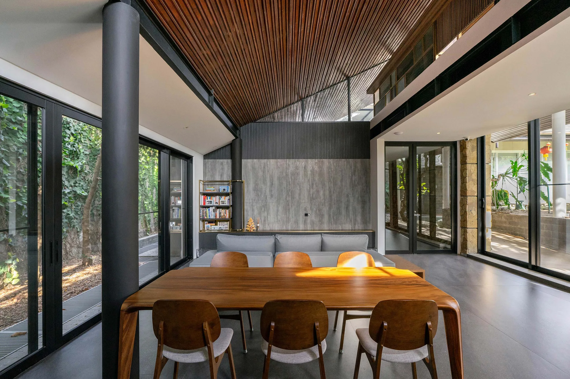 A modern dining room features a wood table and chairs under a slatted timber ceiling, with floor-to-ceiling glass doors showing garden greenery.