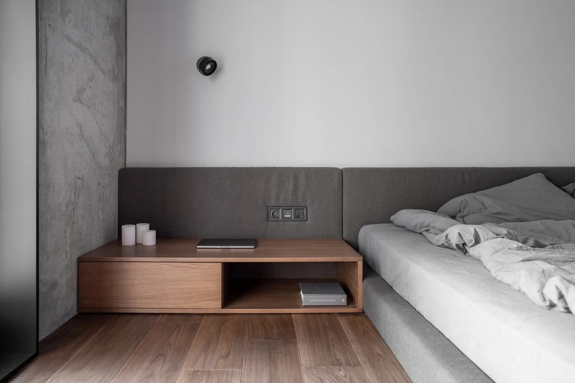 A low-profile grey bed features an extended upholstered headboard and a built-in wooden nightstand against a white wall and concrete panel.