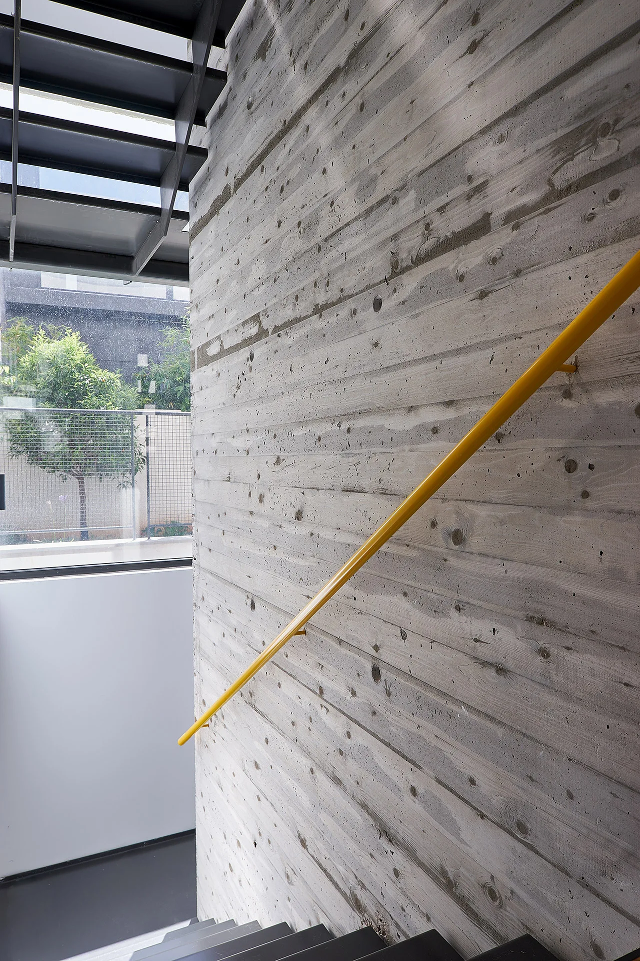 A minimalist staircase features a textured board-formed concrete wall, a bright yellow metal handrail, and dark metal stair treads.