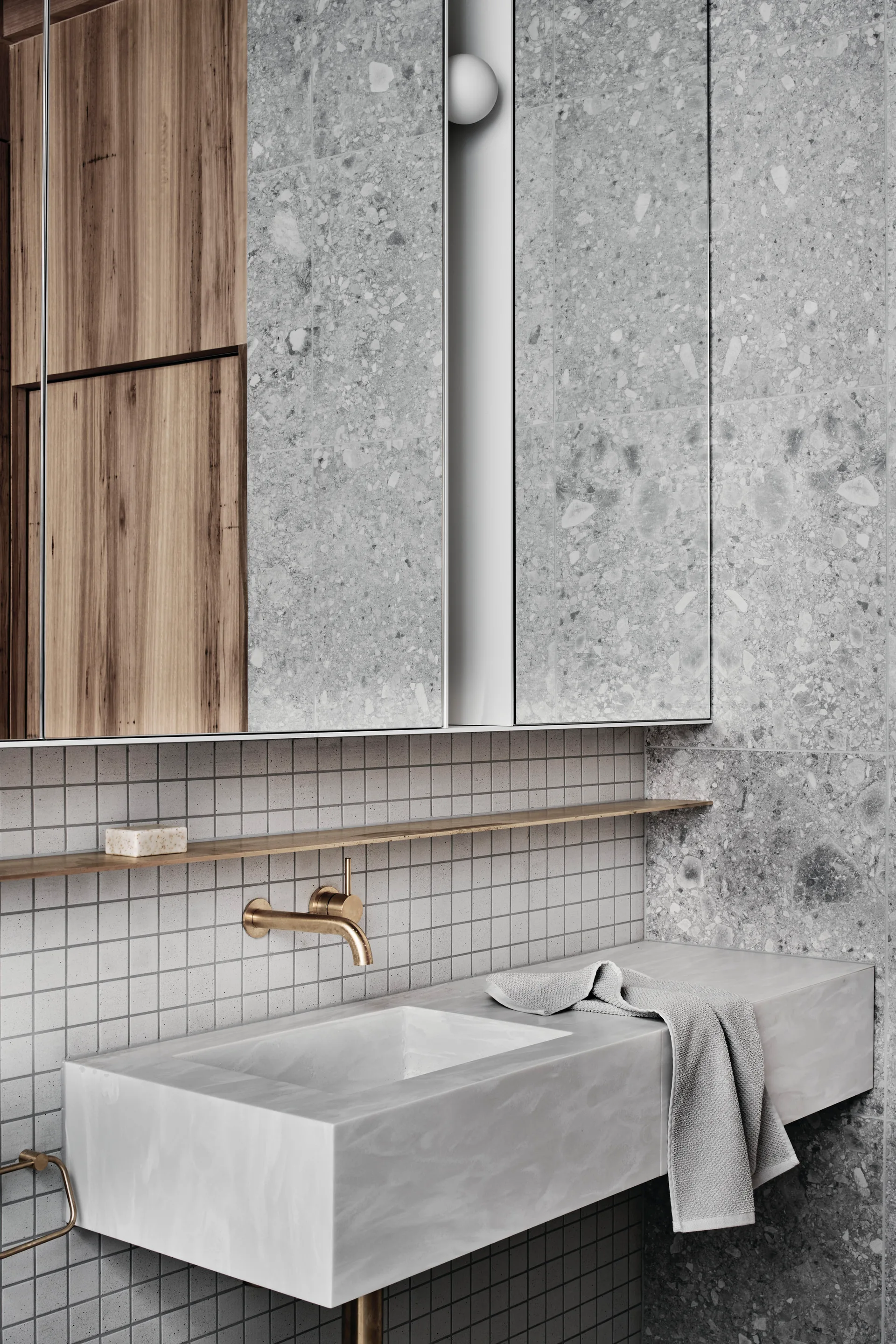 A grey stone bathroom vanity features a gold wall-mounted faucet, a thin metal shelf, square white wall tiles, and terrazzo panels.