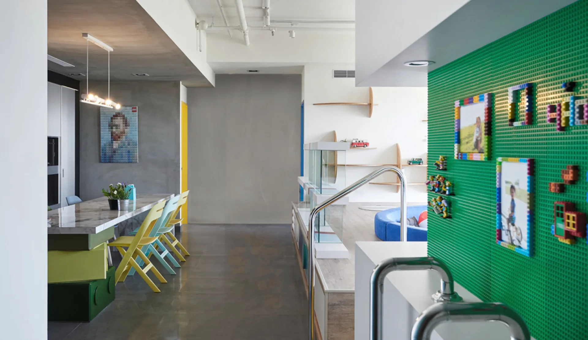 A modern home interior features a marble dining table with colorful chairs next to a raised platform living area and a bright green Lego wall.