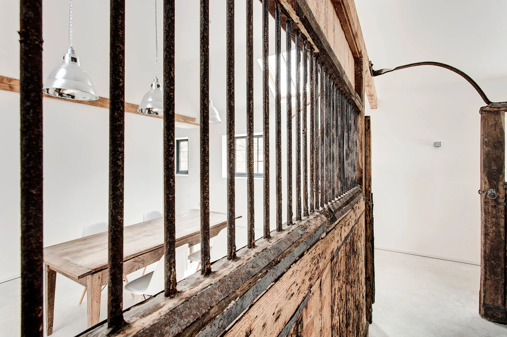 A rustic wood and iron stable door partitions a dining room featuring a long wooden table, white chairs, and silver pendant lights.