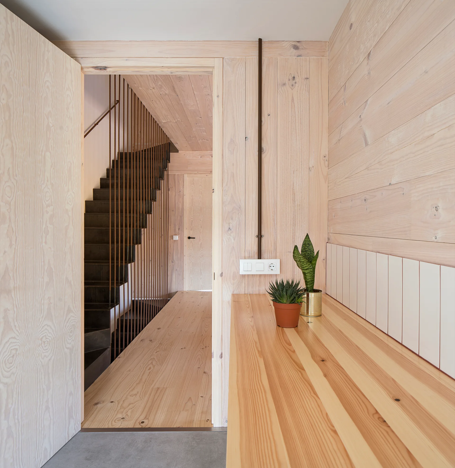 A modern hallway lined with light wood panels features a wooden counter and a doorway looking in on a dark staircase with vertical metal railings.