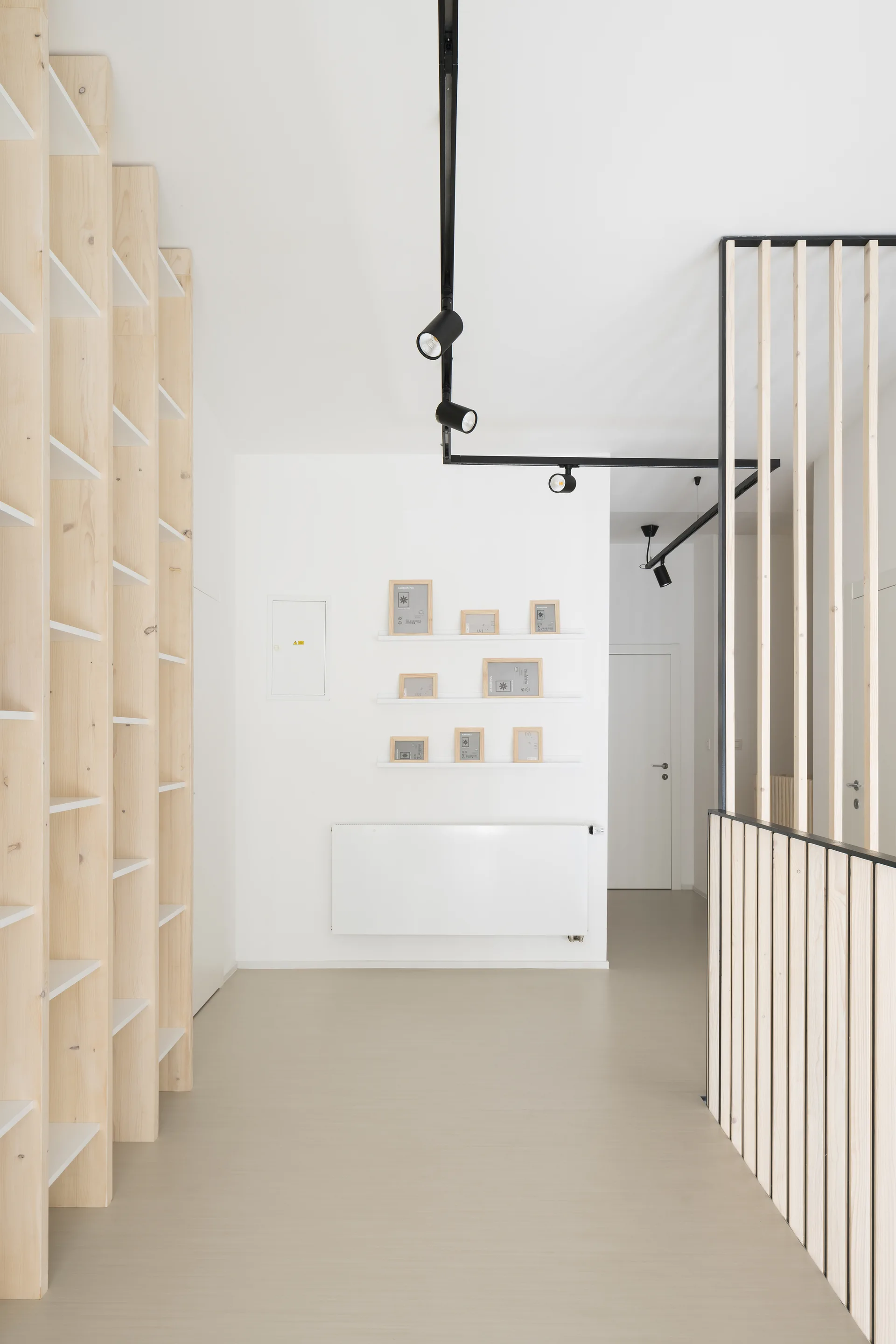 A minimalist white hallway features tall light wood shelving on one side, a vertical wood slat partition on the other, and black track lighting overhead.