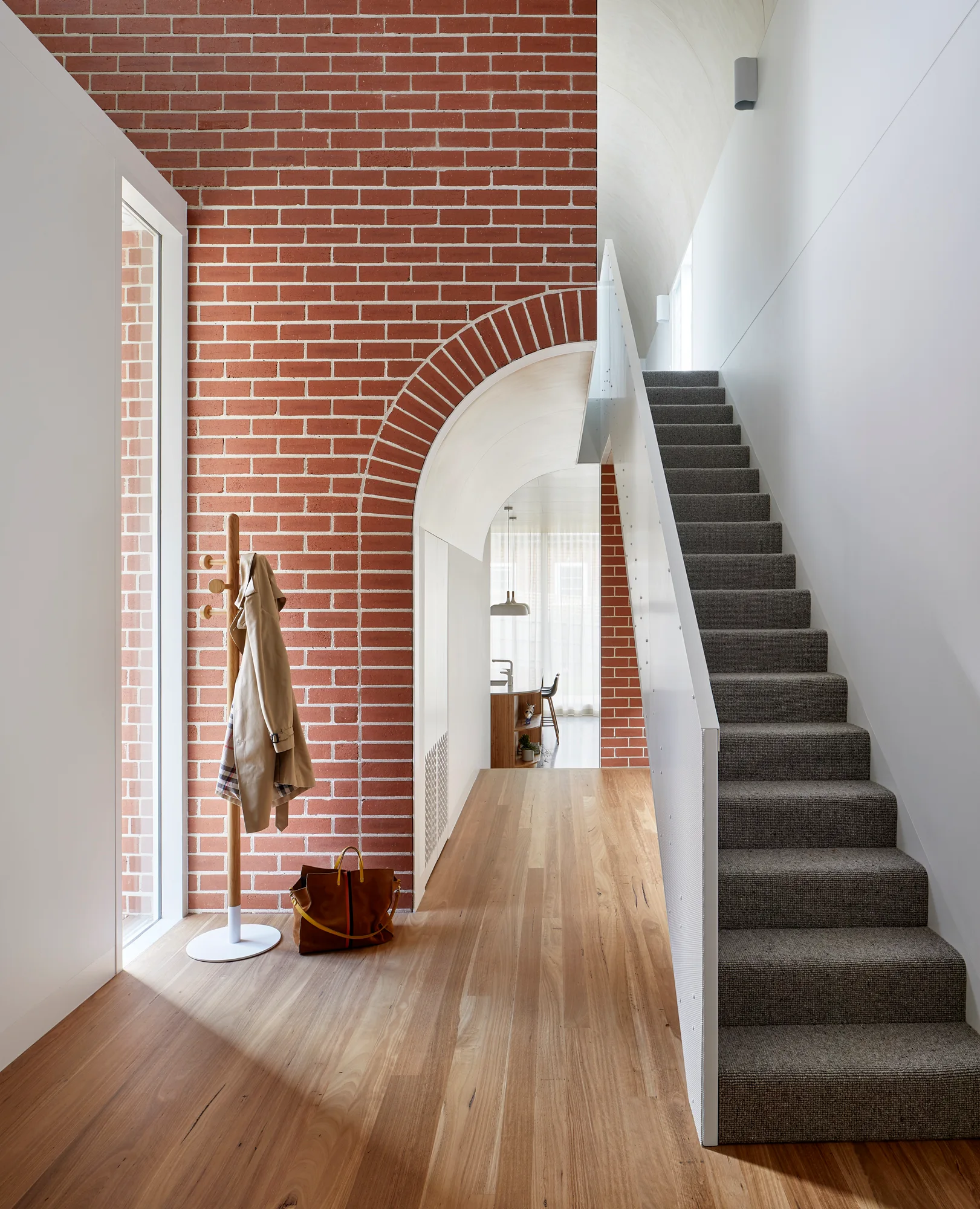 A modern entryway featuring a prominent red brick archway, light wood flooring, a wooden coat rack, and a grey carpeted staircase.