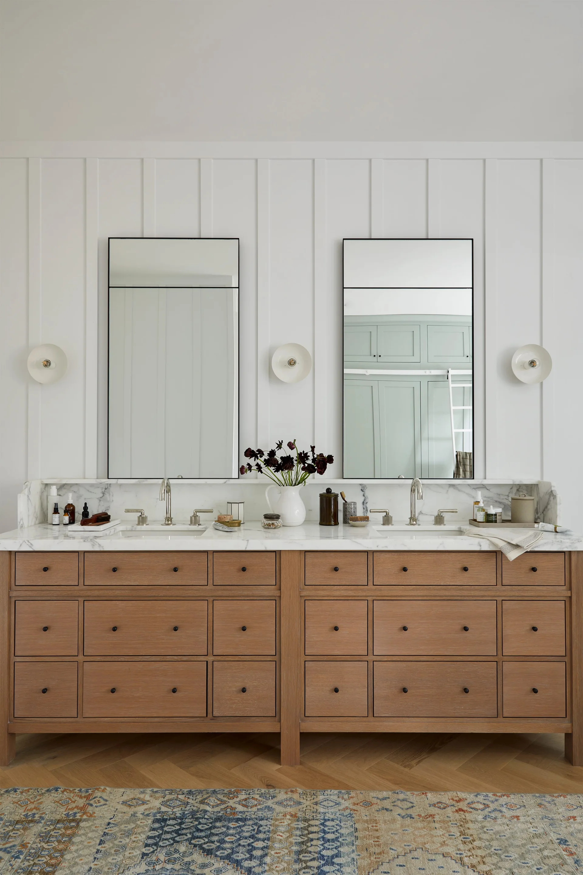 A light wood double vanity features a white marble countertop, two tall black-framed mirrors, and white vertical wall paneling.