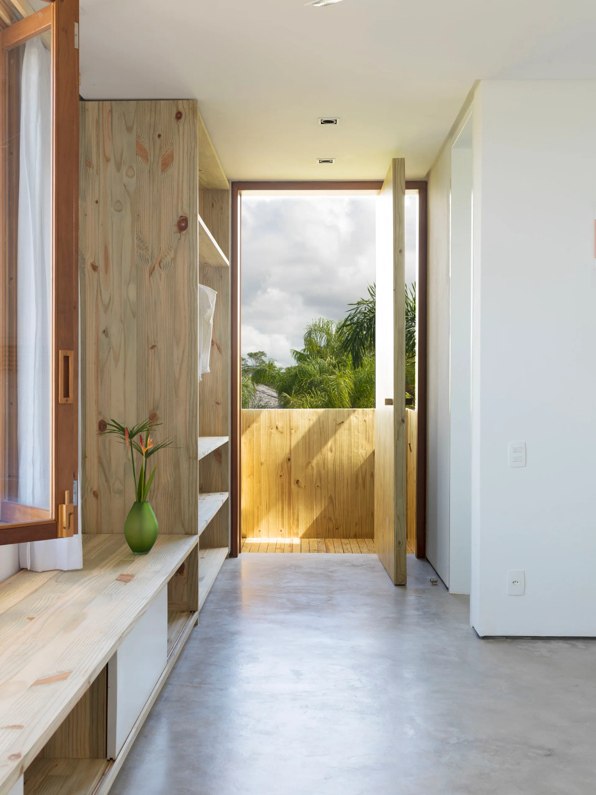 A minimalist dressing room features light pine wood built-in shelving, concrete flooring, and an open door leading to a wood-paneled balcony.