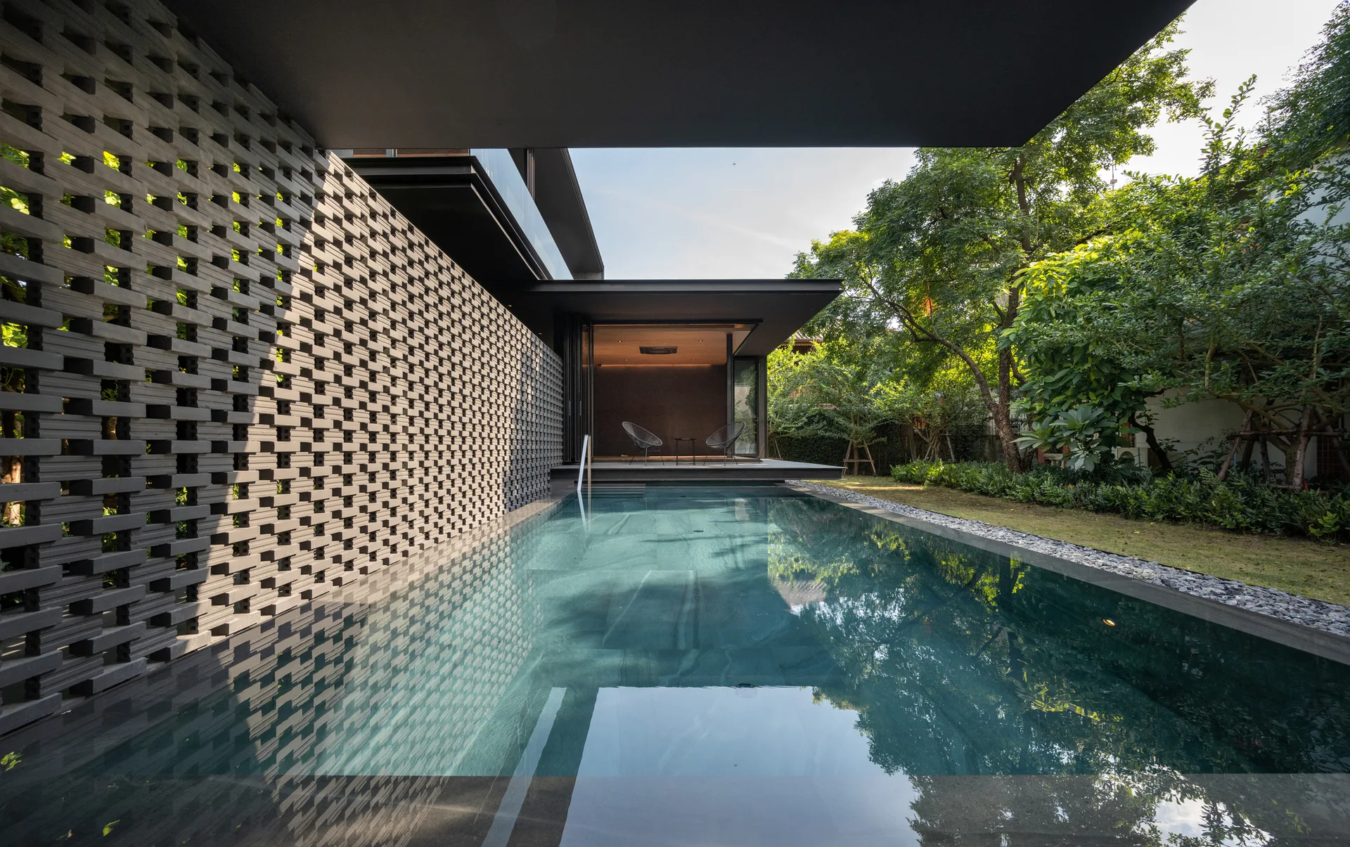 A rectangular swimming pool is flanked by a grey perforated brick wall and a green garden under a dark architectural roof overhang.