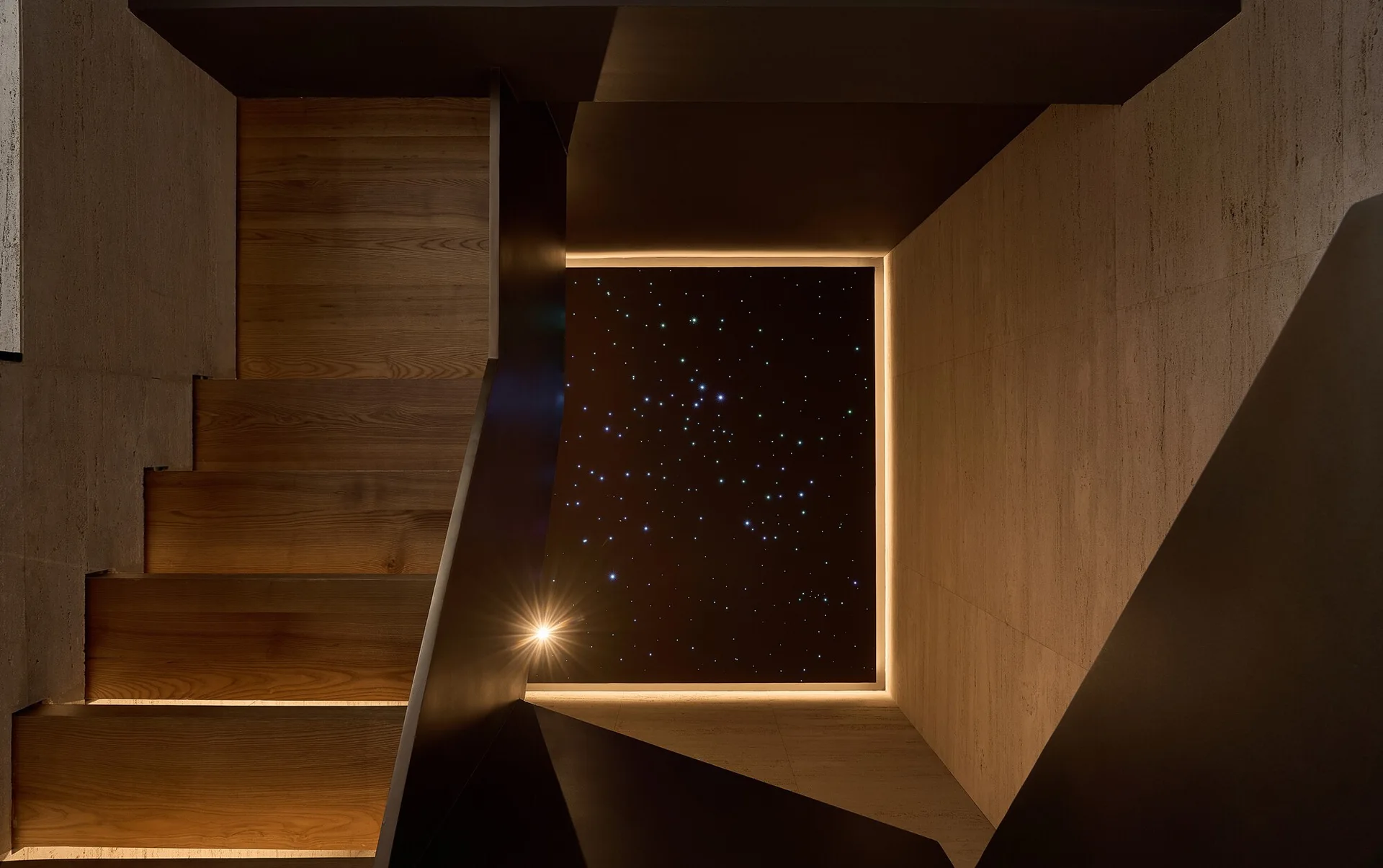 A modern wooden staircase winding upward beside textured stone walls toward a dark ceiling panel illuminated with star-like fiber optic lights.