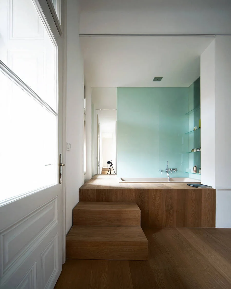 A minimalist bathroom featuring a recessed bathtub set into a raised wood platform with integrated steps, set against a pale mint green glass wall.