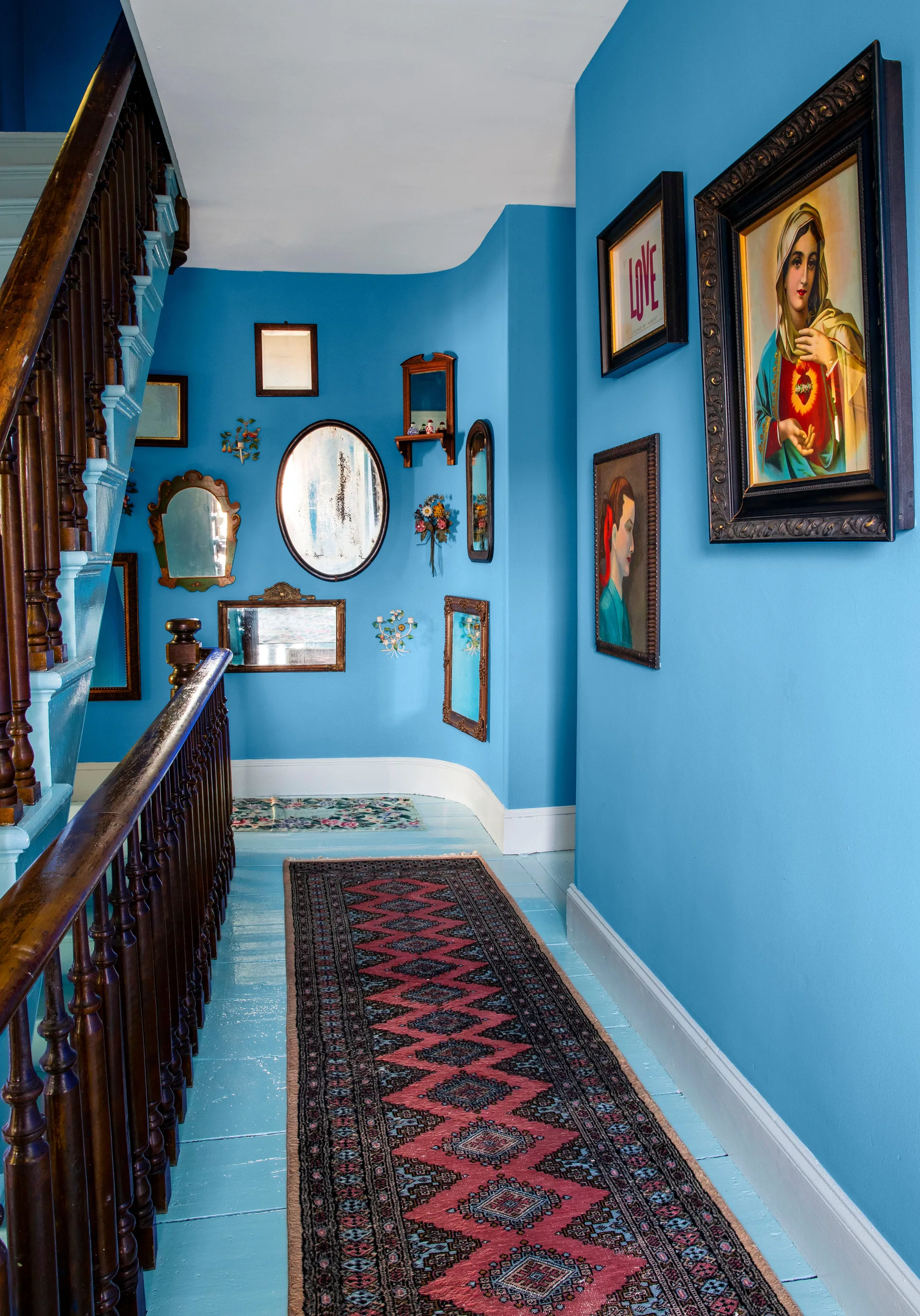 A bright blue hallway features painted blue floorboards, a dark wood staircase banister, a patterned runner rug, and a wall of eclectic mirrors.