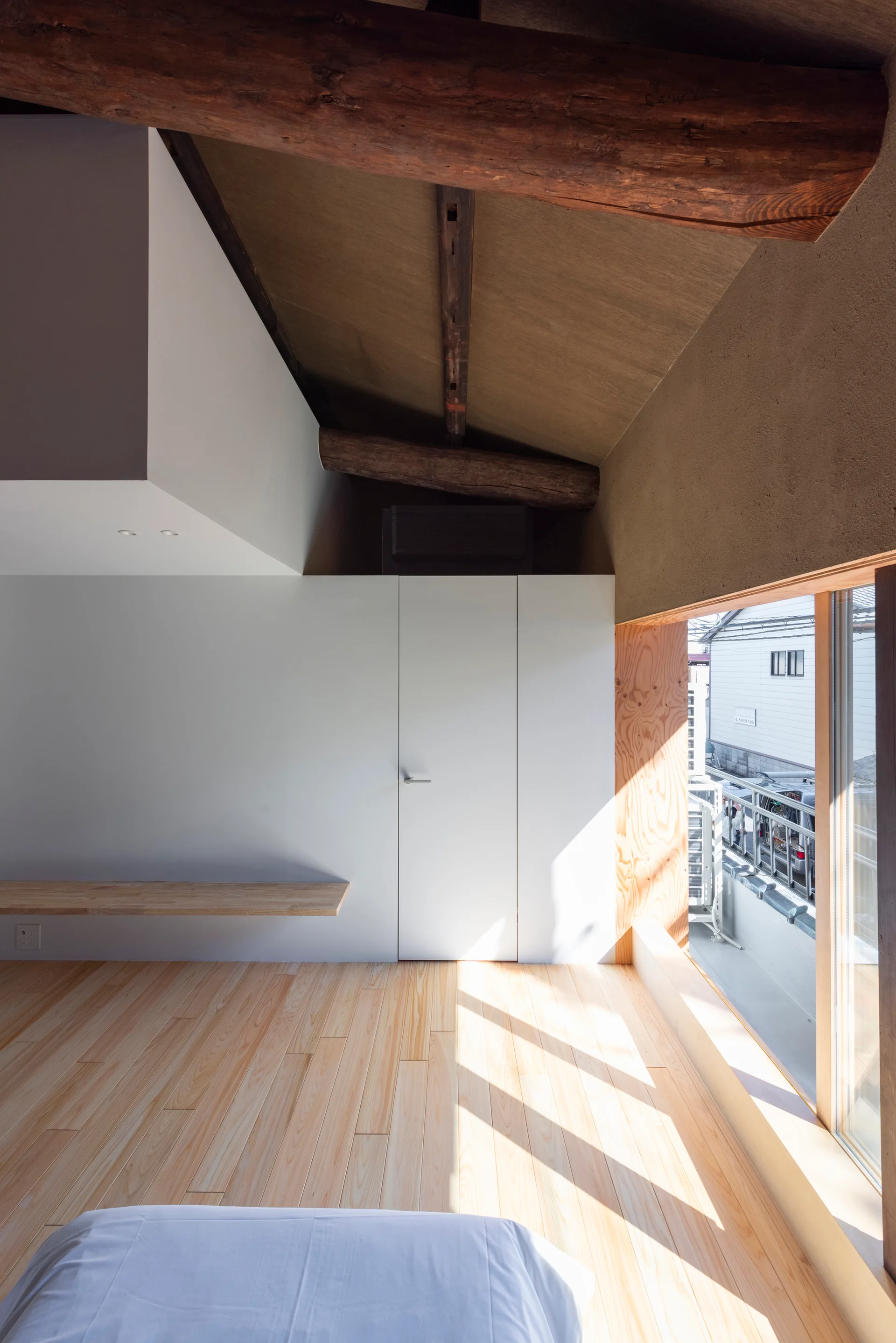 A minimalist bedroom featuring light wood flooring, rustic exposed ceiling beams, flat white closet doors, and a low plywood-framed window.