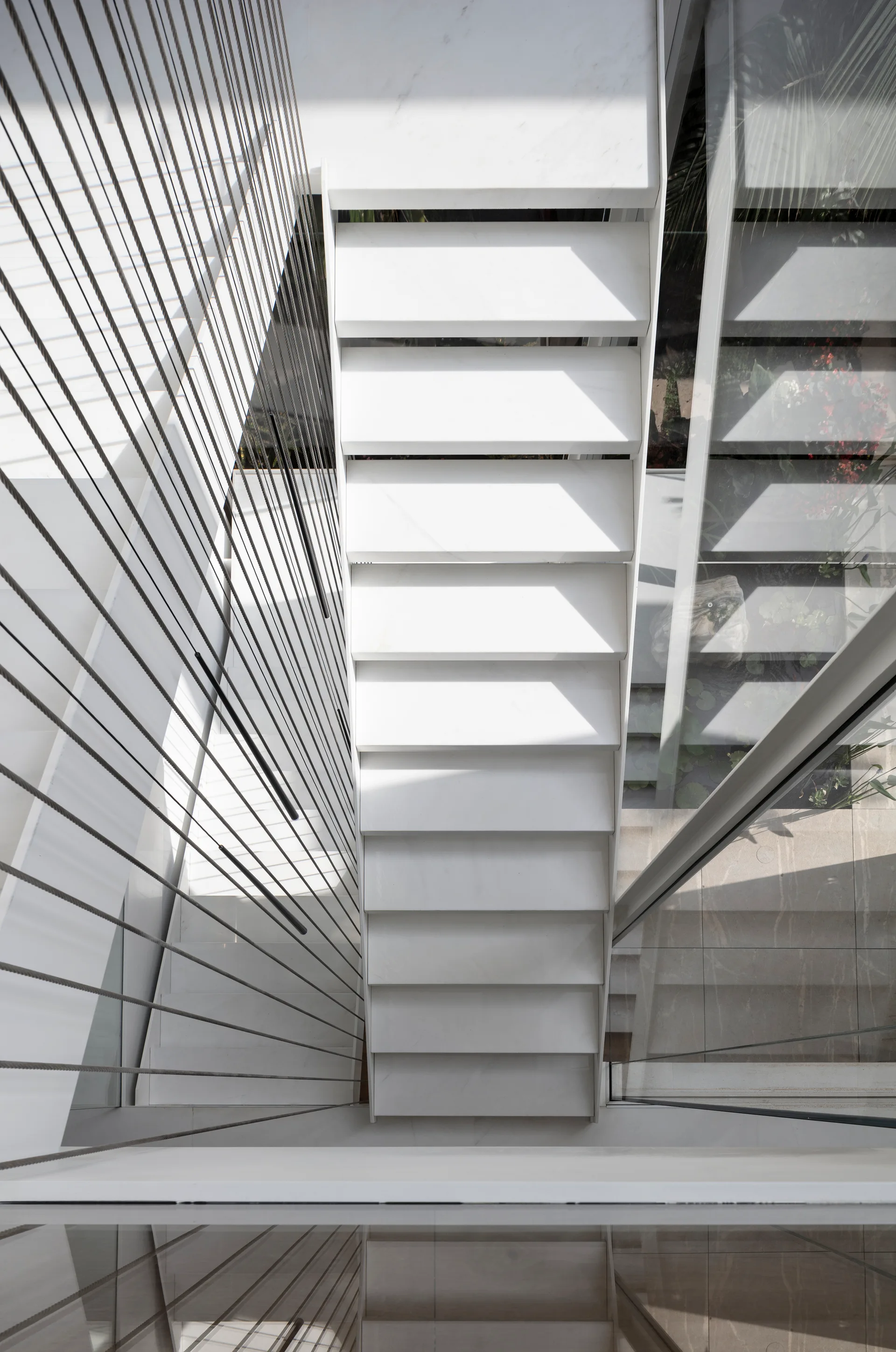 Looking down a modern white floating staircase enclosed by vertical metal tension cables and a clear glass wall under bright natural light.